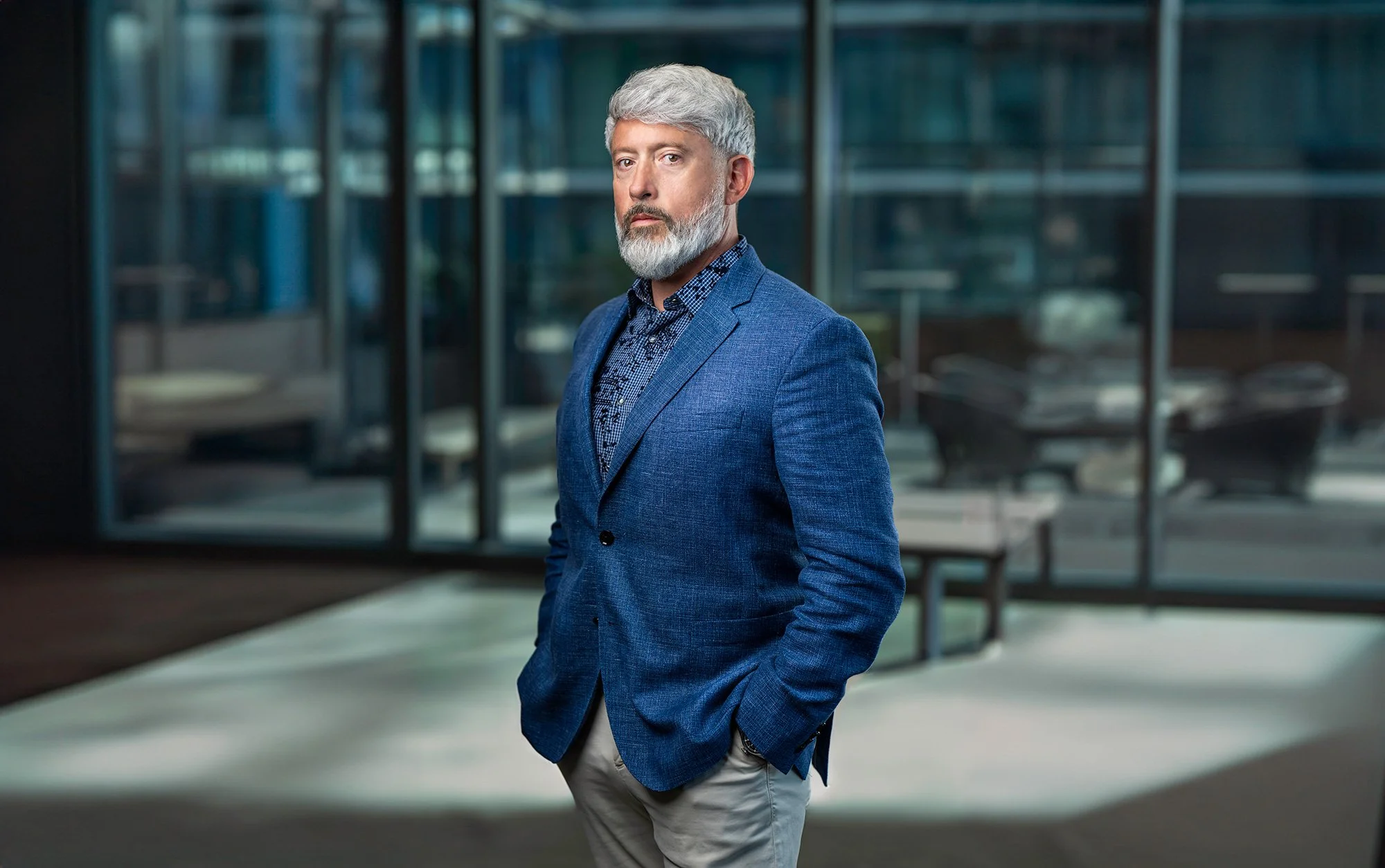 A middle-aged man with gray hair and beard wearing a blue blazer and light-colored pants, standing outside in front of a modern glass building.