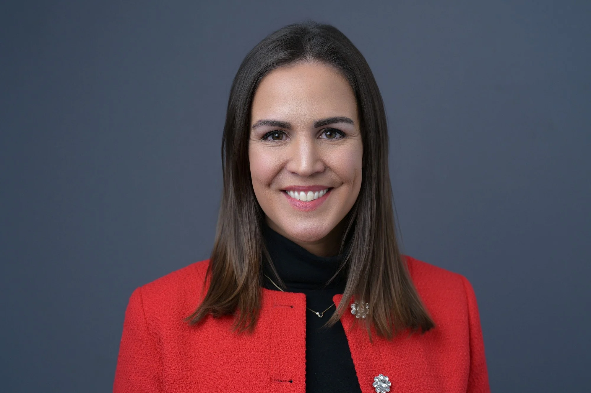 Portrait of a woman with long brown hair, smiling, wearing a red blazer with decorative buttons and a black turtleneck, against a gray background.