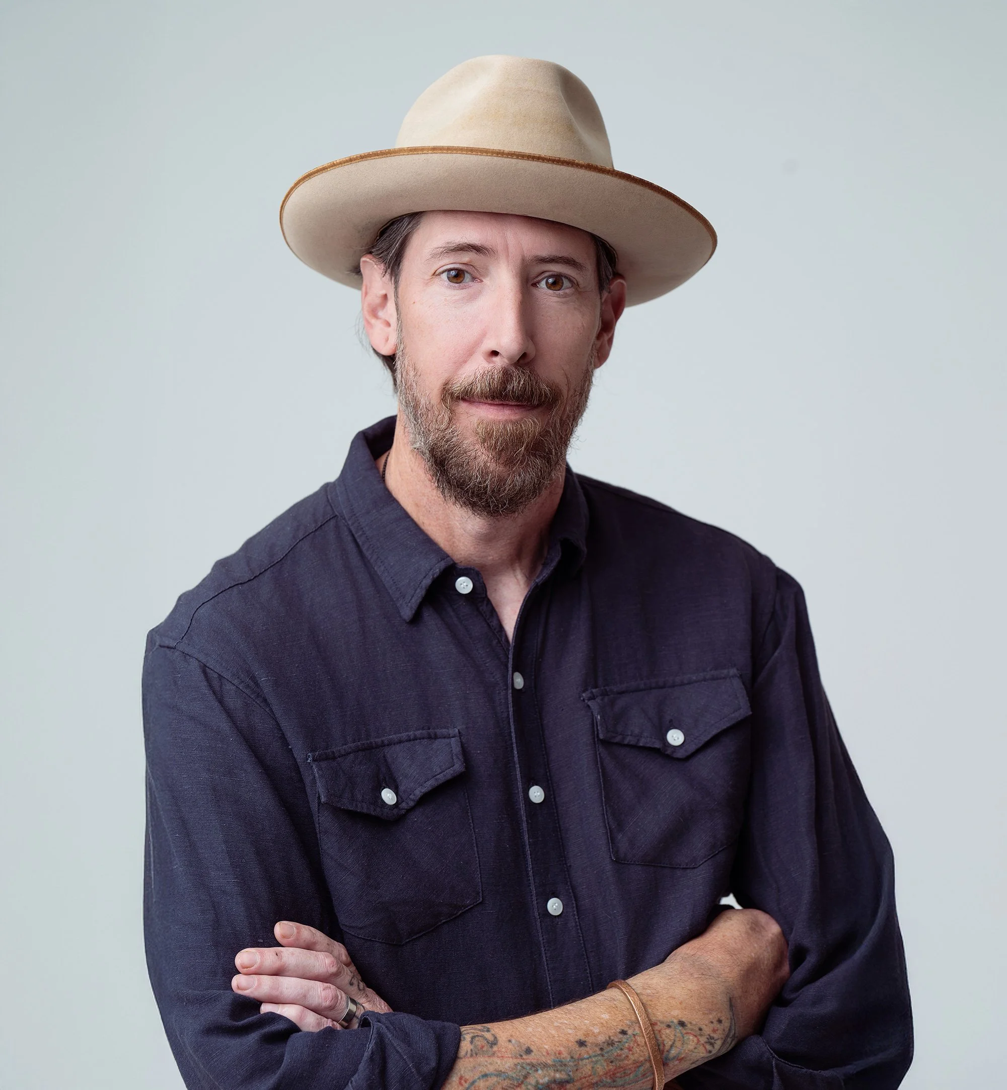 A man with a beard wearing a beige wide-brimmed hat and a dark button-up shirt, standing against a light background.