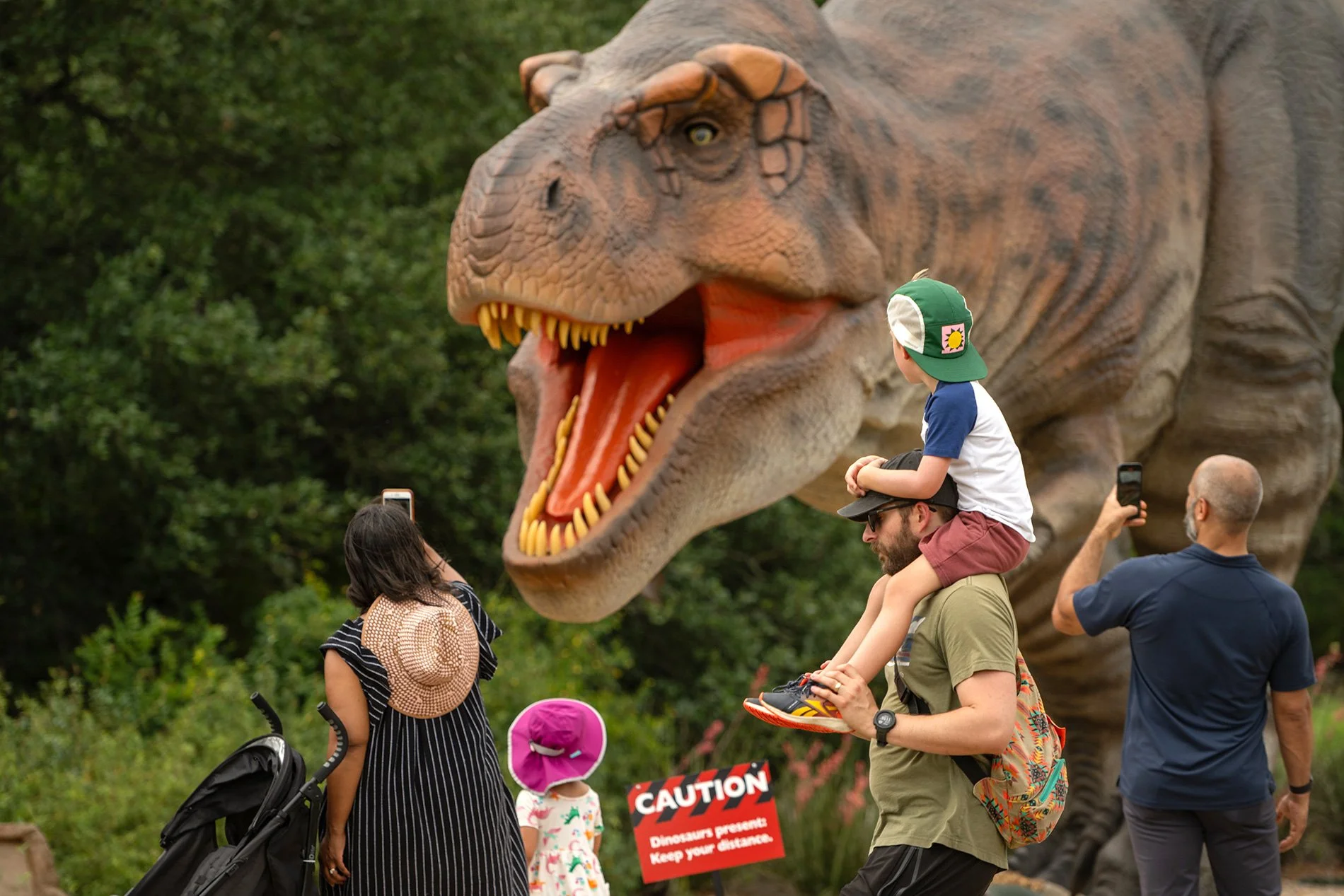 People taking photos of a large dinosaur statue in an outdoor setting, with a warning sign about keeping distance from dinosaurs.