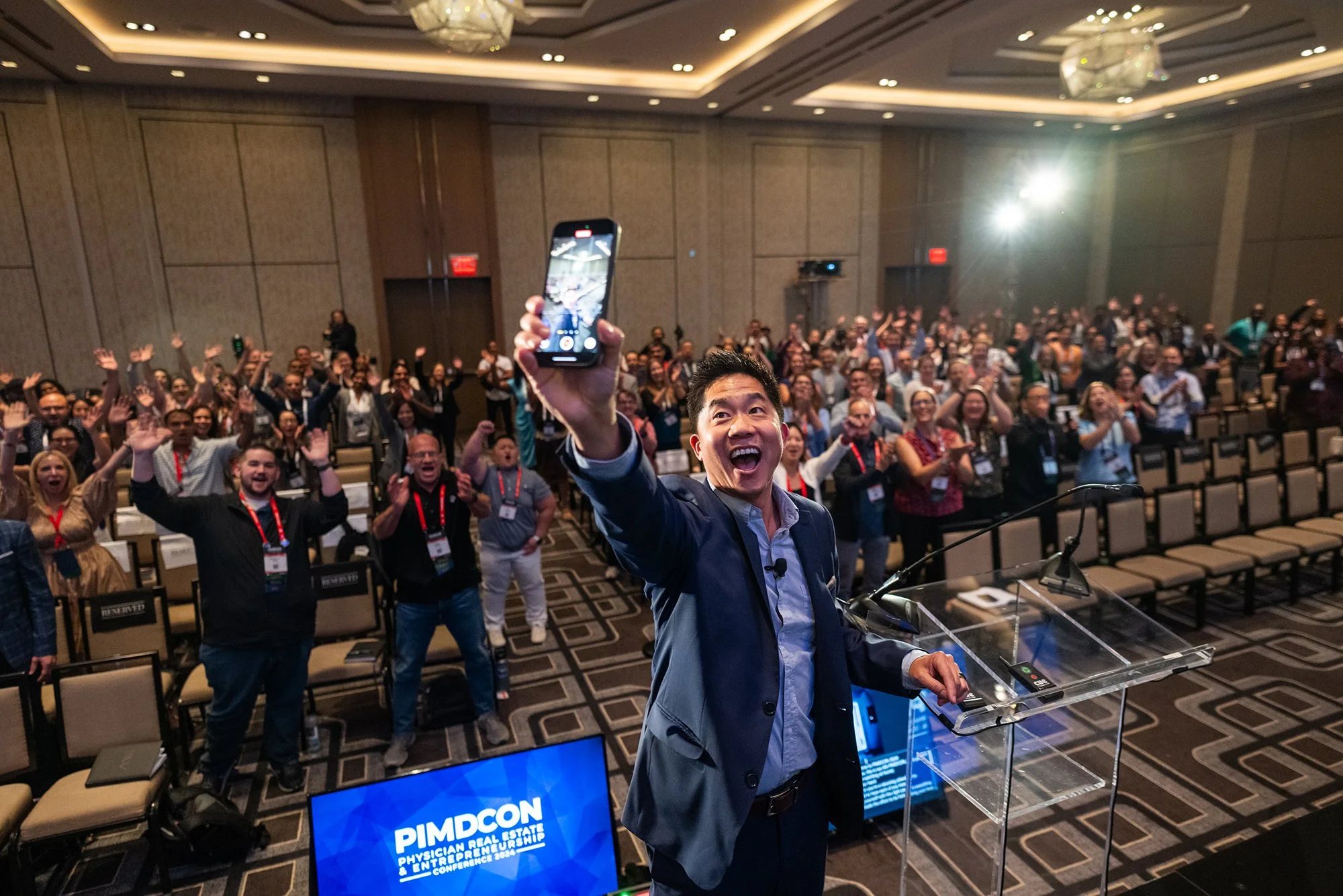 A man in a suit standing at a podium, taking a selfie with a large group of smiling, cheering people at a conference or event, with a PIMDCON sign on a screen in front captured by tico mendoza corporate event photographer austin tx