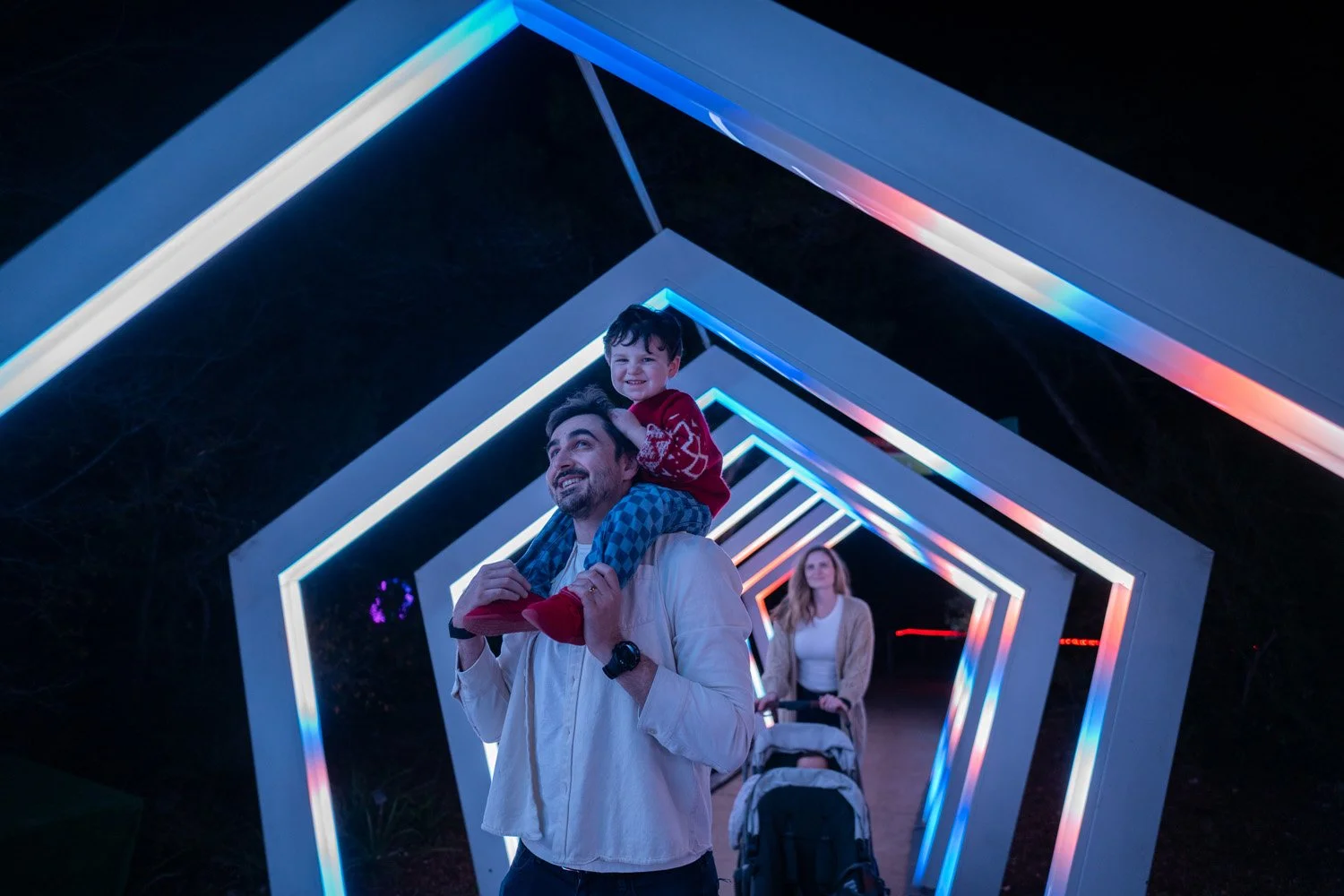 A man carrying a young boy on his shoulders, with a woman and stroller in the background, walking through illuminated geometric tunnel at night.