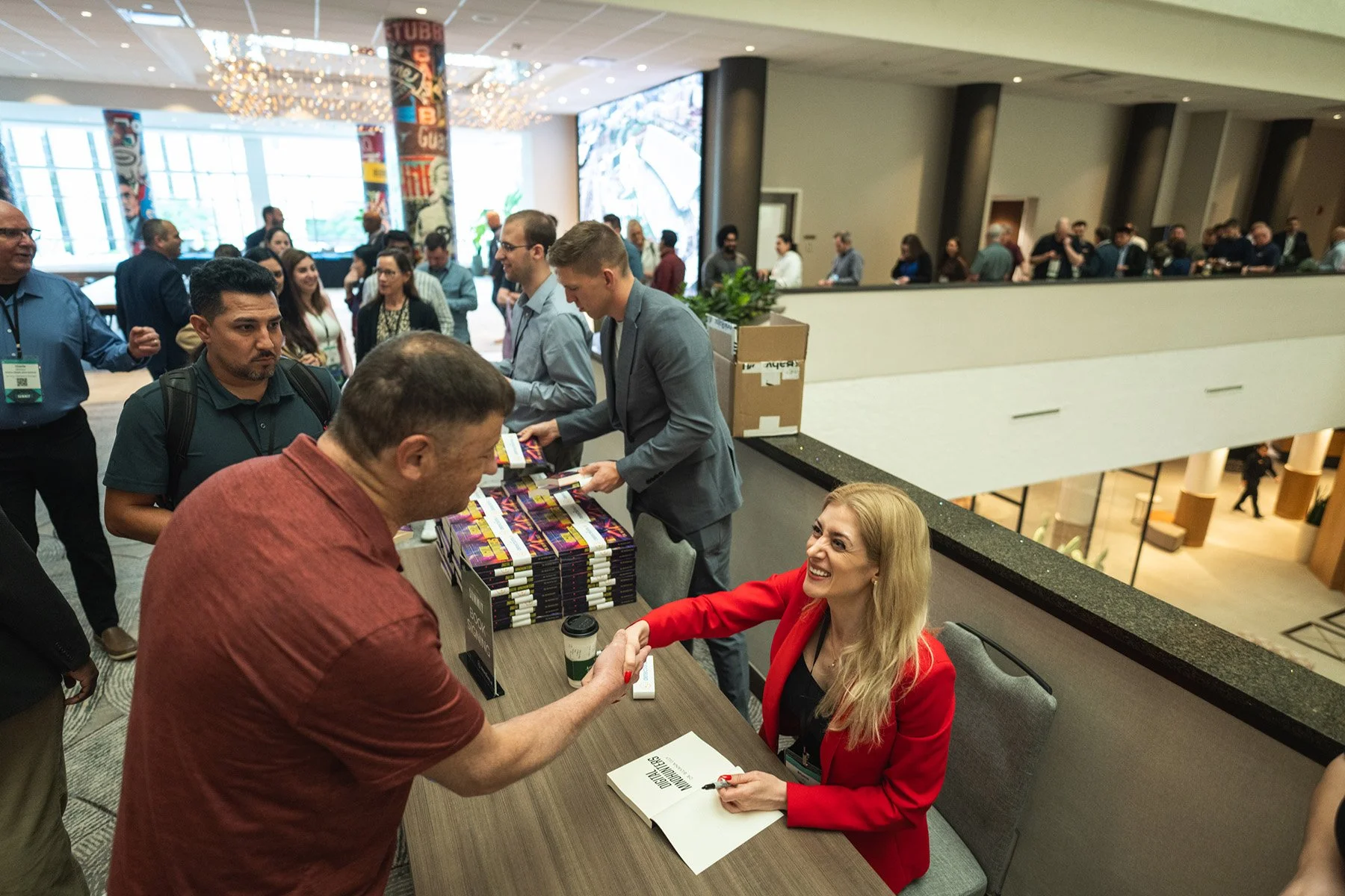 Austin corporate event photography capturing a speaker in a red blazer signing books and shaking hands with an attendee in a large conference hall.