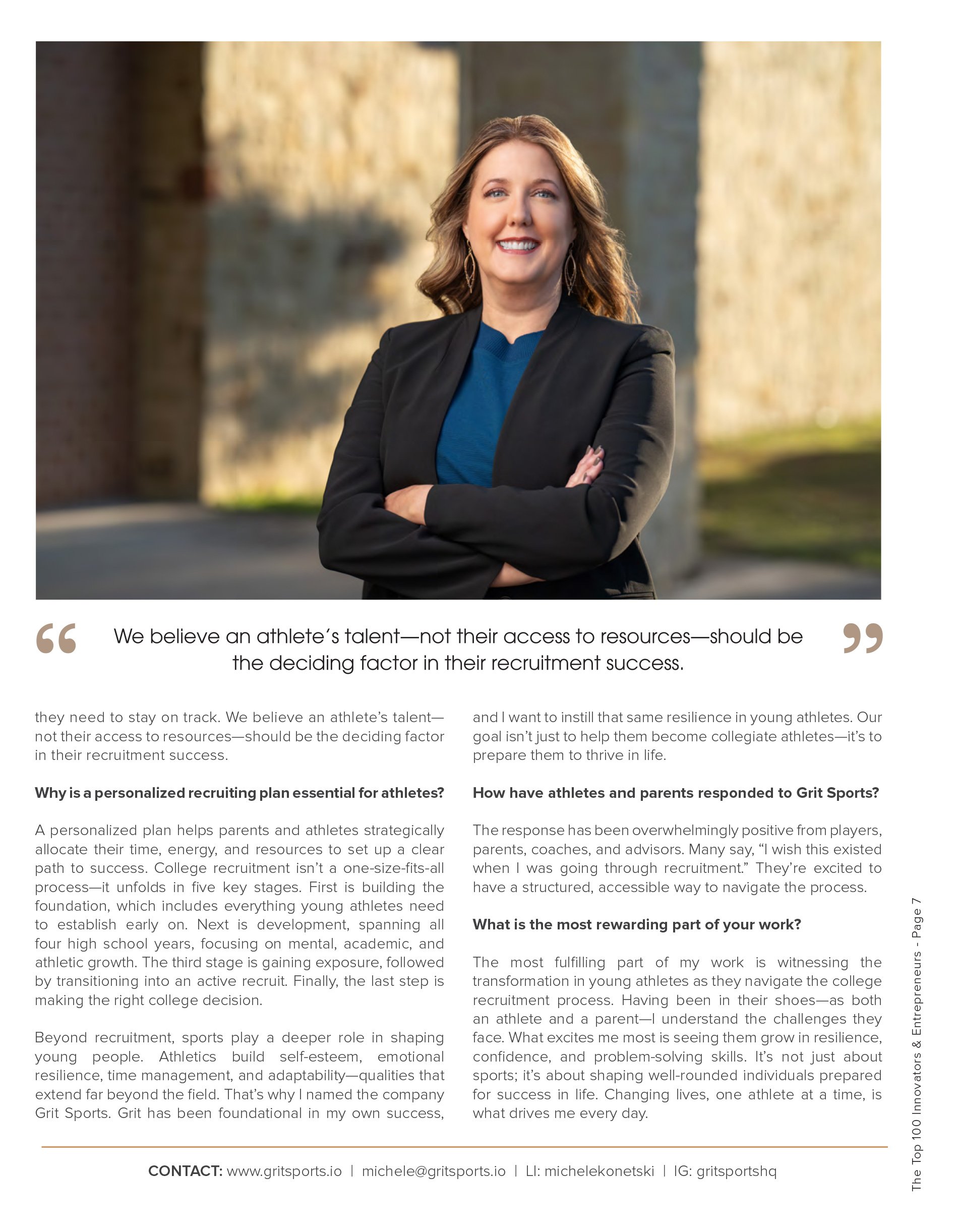 A confident woman outdoors with crossed arms, wearing a black blazer and blue top, smiling at the camera. Behind her is a stone wall and grassy area.