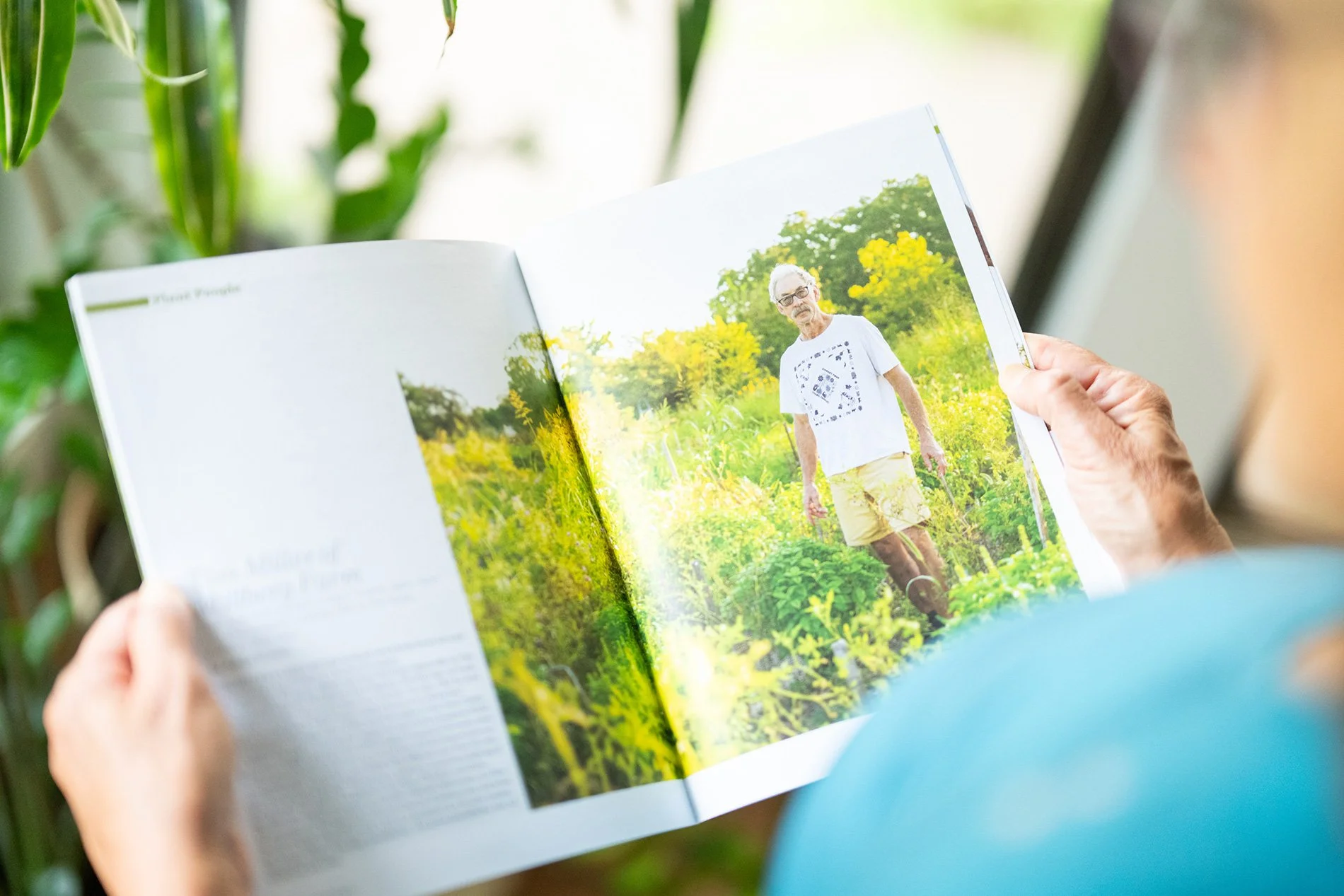 Person holding an open magazine or book with a photo of an elderly man standing in a lush garden or field with yellow flowers.