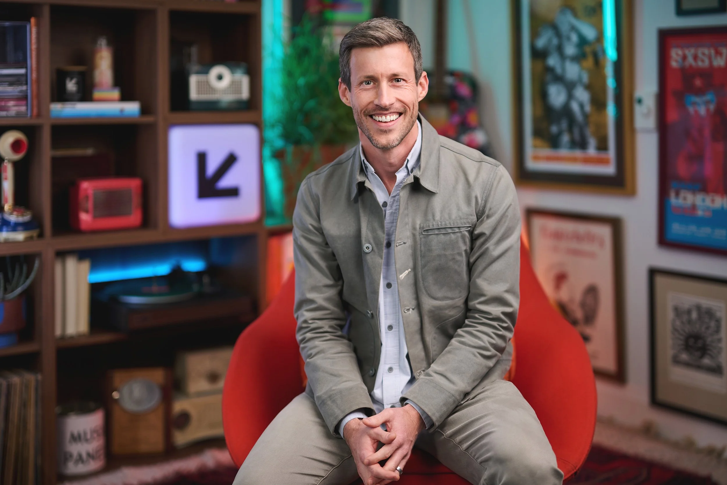 A smiling man with short brown hair and light stubble sits on a red chair in a colorful room. Behind him are shelves with books, decorations, and framed posters on the wall.