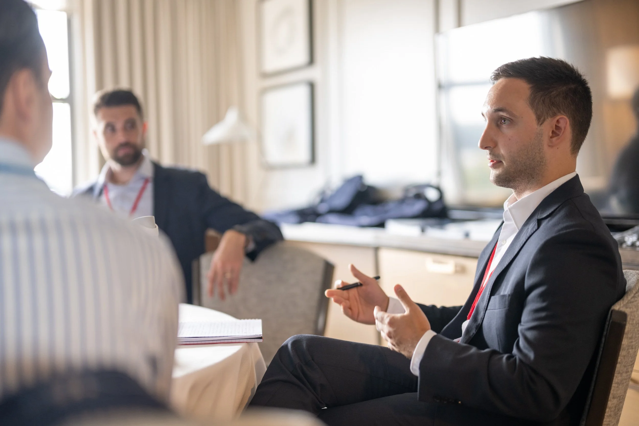 Three men in business attire sitting and talking in a bright, window-lit room, with one man holding a pen.