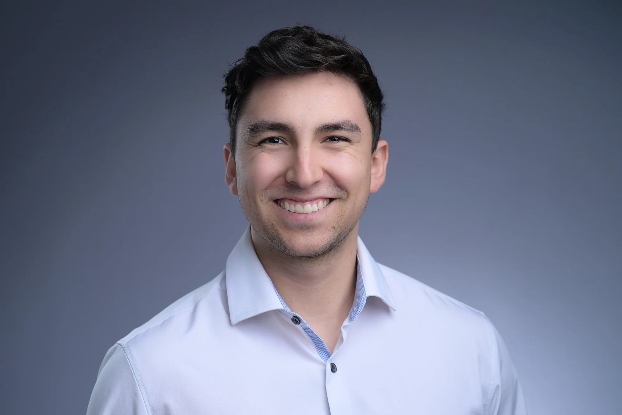 Headshot of a smiling young man with dark hair in a white shirt against a gray background.