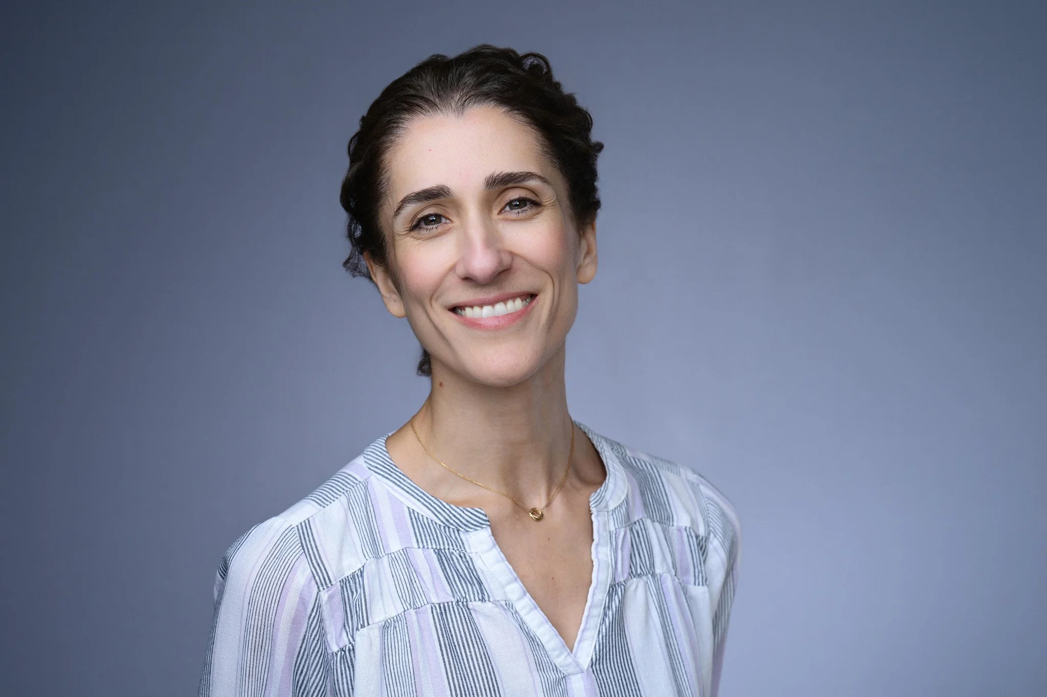A woman smiling with short dark hair, wearing a striped shirt and a gold necklace, standing against a plain blue background.