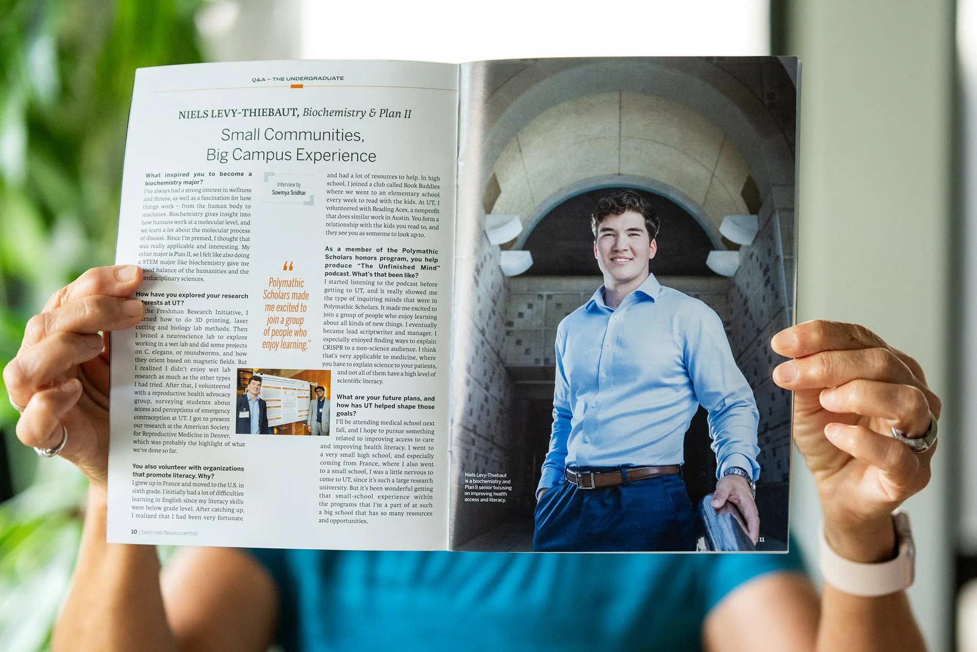 Magazine open to an interview article with a young man in a blue shirt smiling in front of a large stone building. Hands holding the magazine are visible, and the person is wearing a white watch.