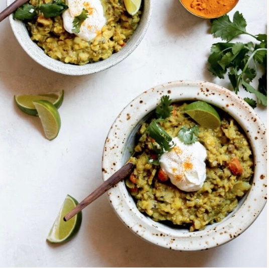 Bowl of yellow lentil soup garnished with cilantro, lime, and a dollop of yogurt. Nearby are lime wedges, cilantro sprigs, and additional bowls of lentil soup on a white surface.