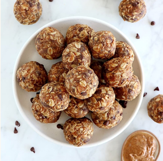Postpartum meal of a white bowl filled with oat and chocolate energy bite balls on a white surface, some scattered around, with a small jar of peanut butter nearby.