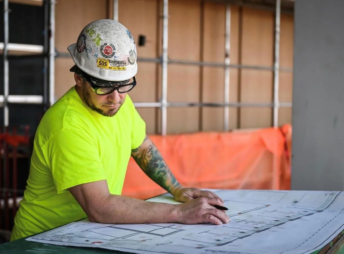 Construction worker wearing a hard hat, safety glasses, and a neon yellow shirt reviewing blueprints at a construction site.