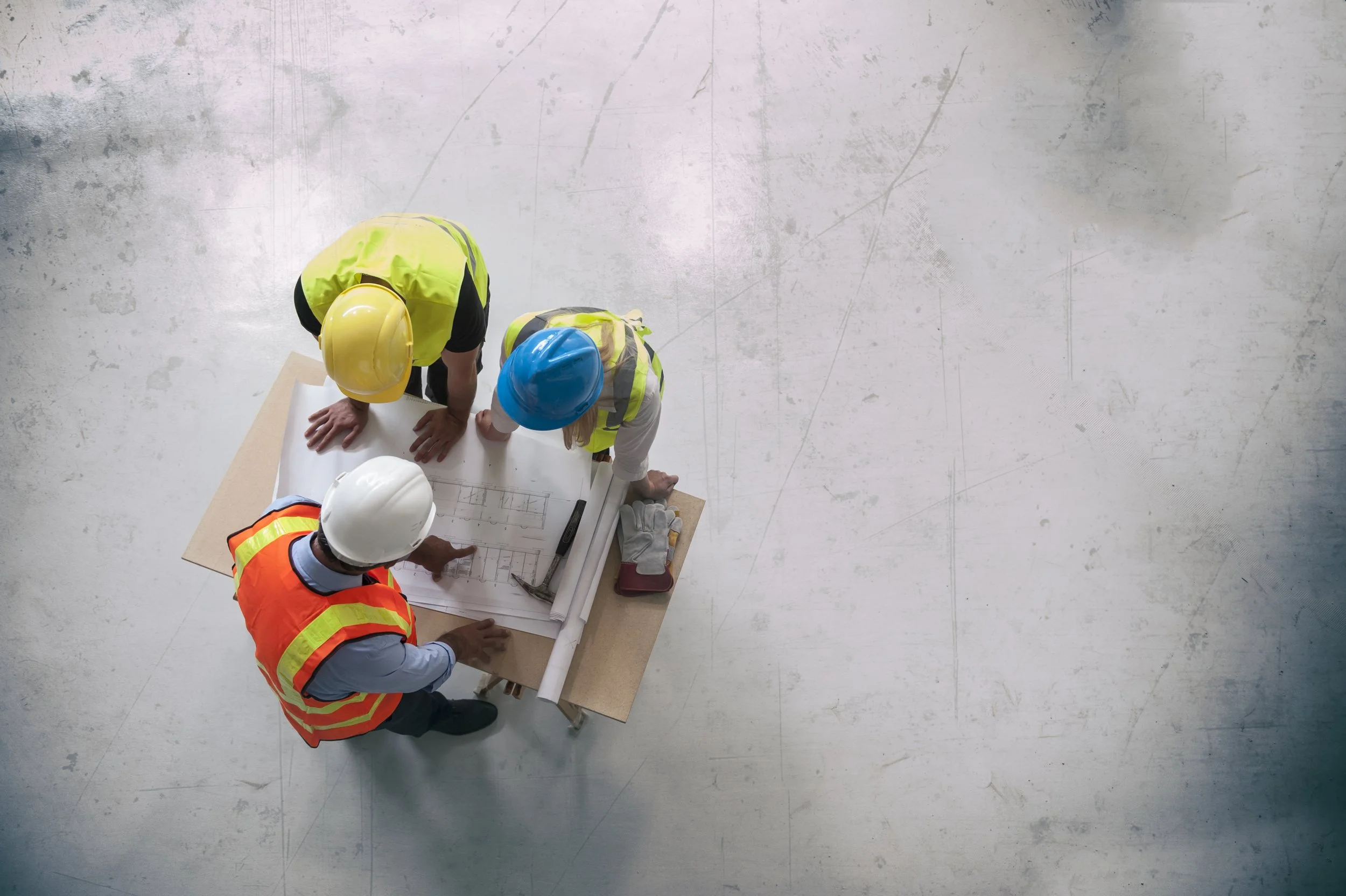 Top view of three construction workers wearing hard hats and safety vests, reviewing architectural blueprints on a table.