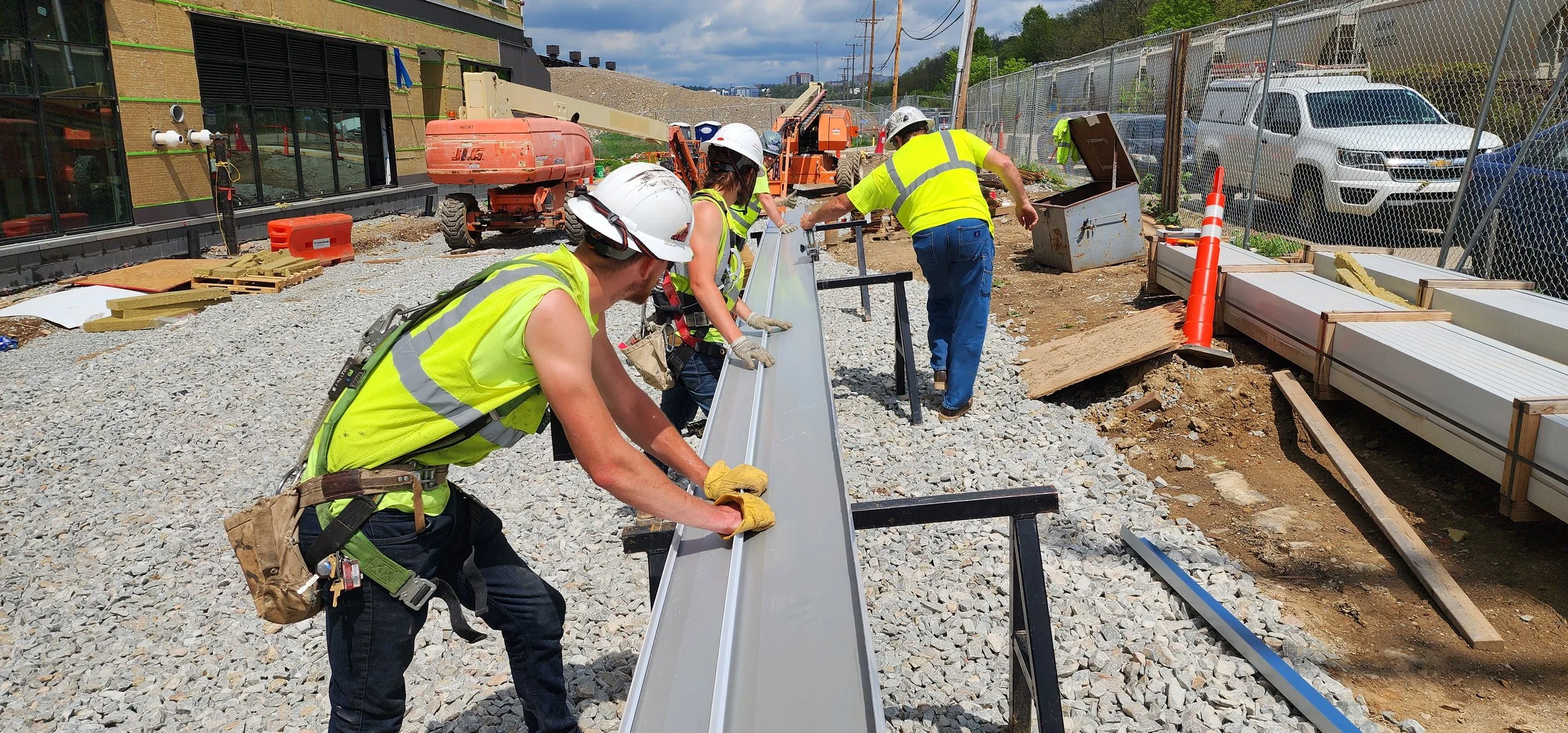 Construction workers installing a metal beam at a construction site with gravel ground, construction tools, and vehicles in the background.