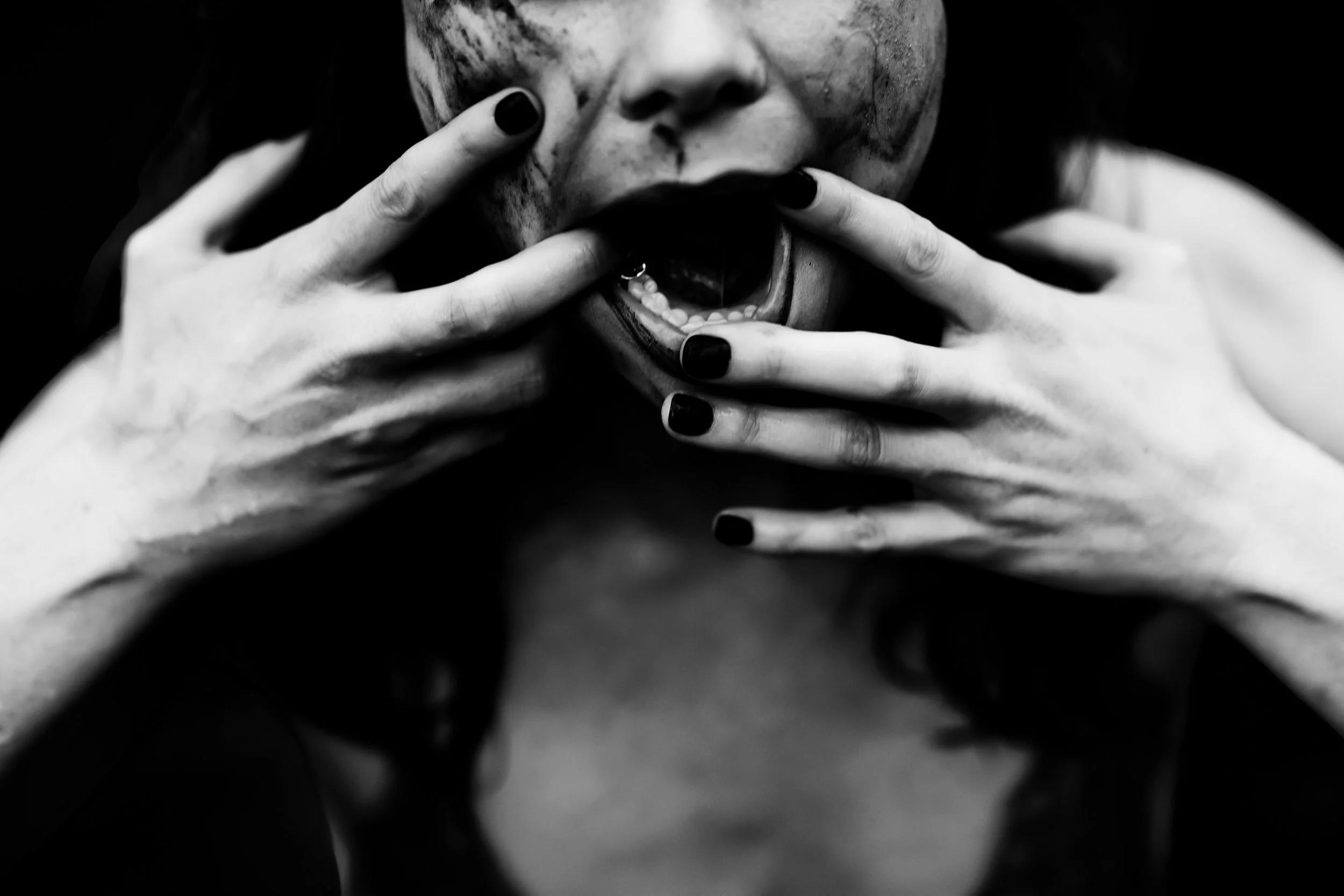Black and white close-up of a person with a mask or face paint, holding their fingers near their mouth, with dark nail polish visible.