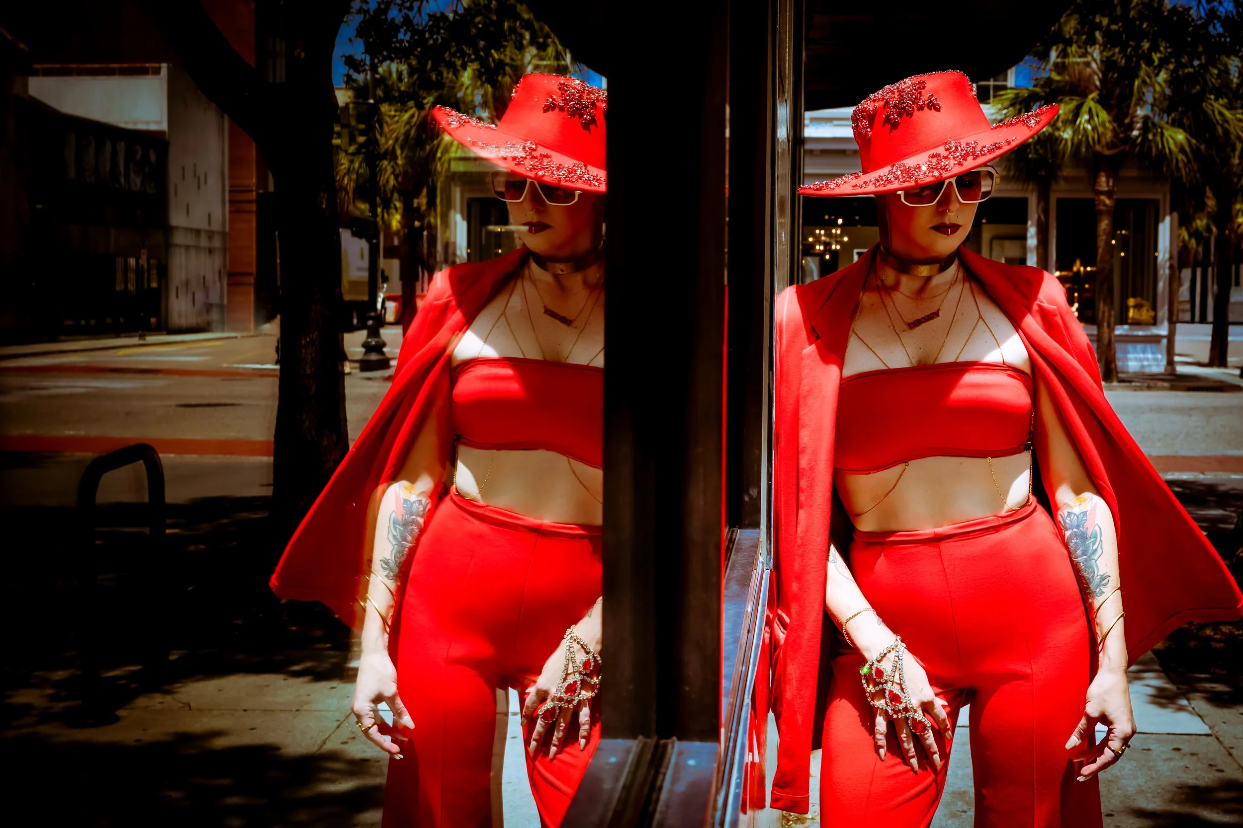 A woman in a red outfit with a wide-brimmed red hat decorated with flowers, sunglasses, and dark lipstick, standing outdoors and looking at her reflection in a mirror.
