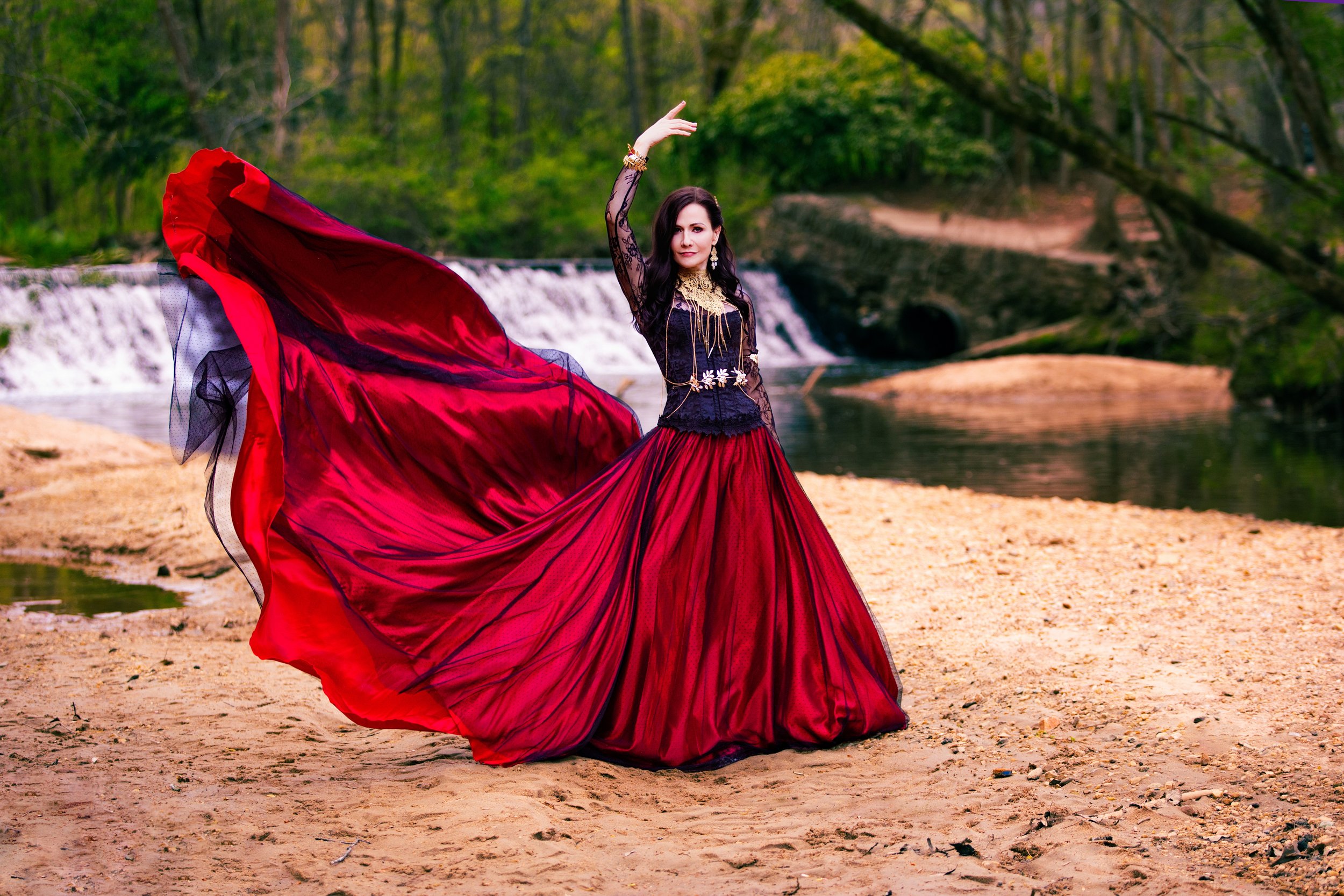 Woman in a long flowing red skirt and black lace top dancing near a creek in a forested area.