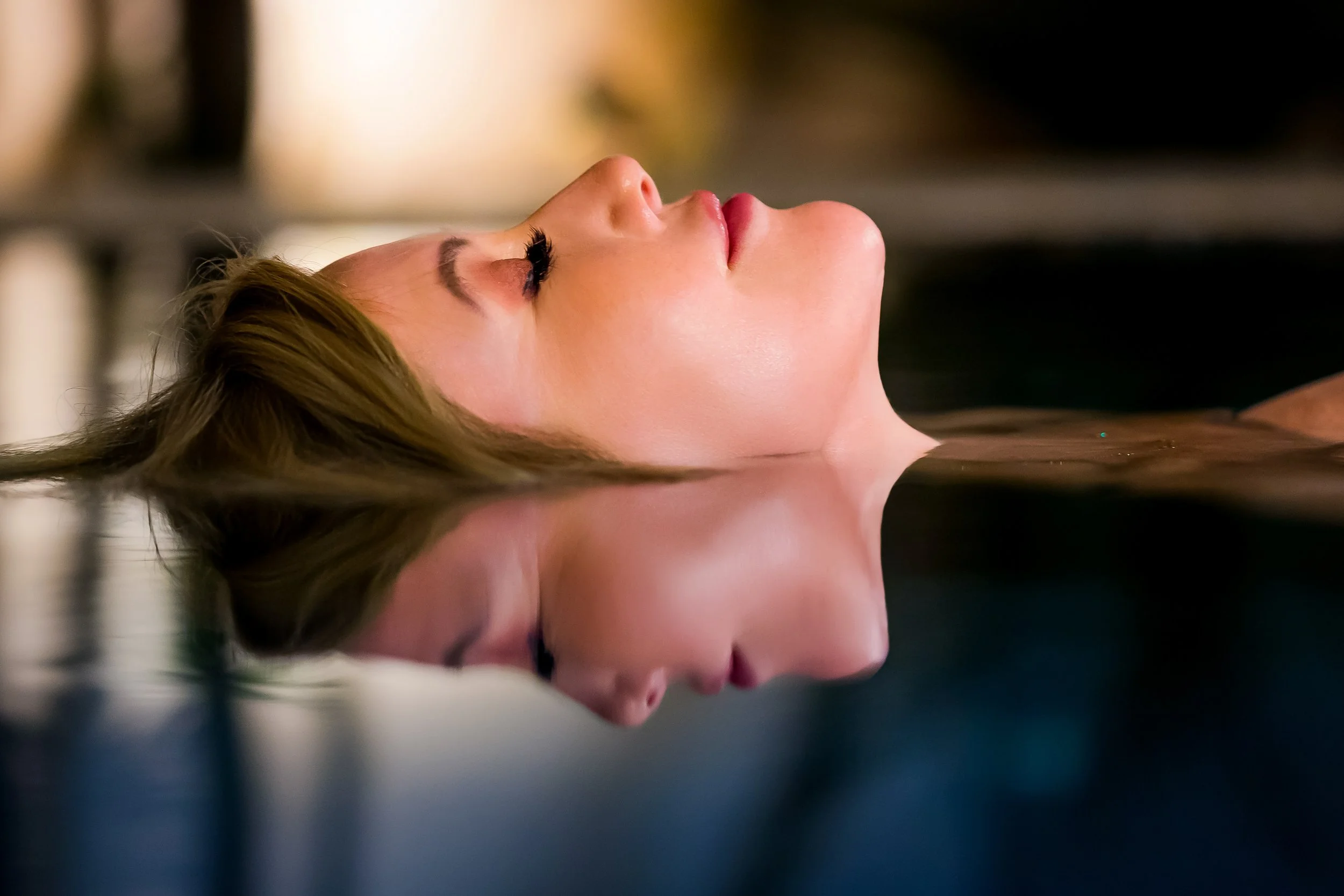 A woman with long hair lying down with her eyes closed, reflected in a pool of water.