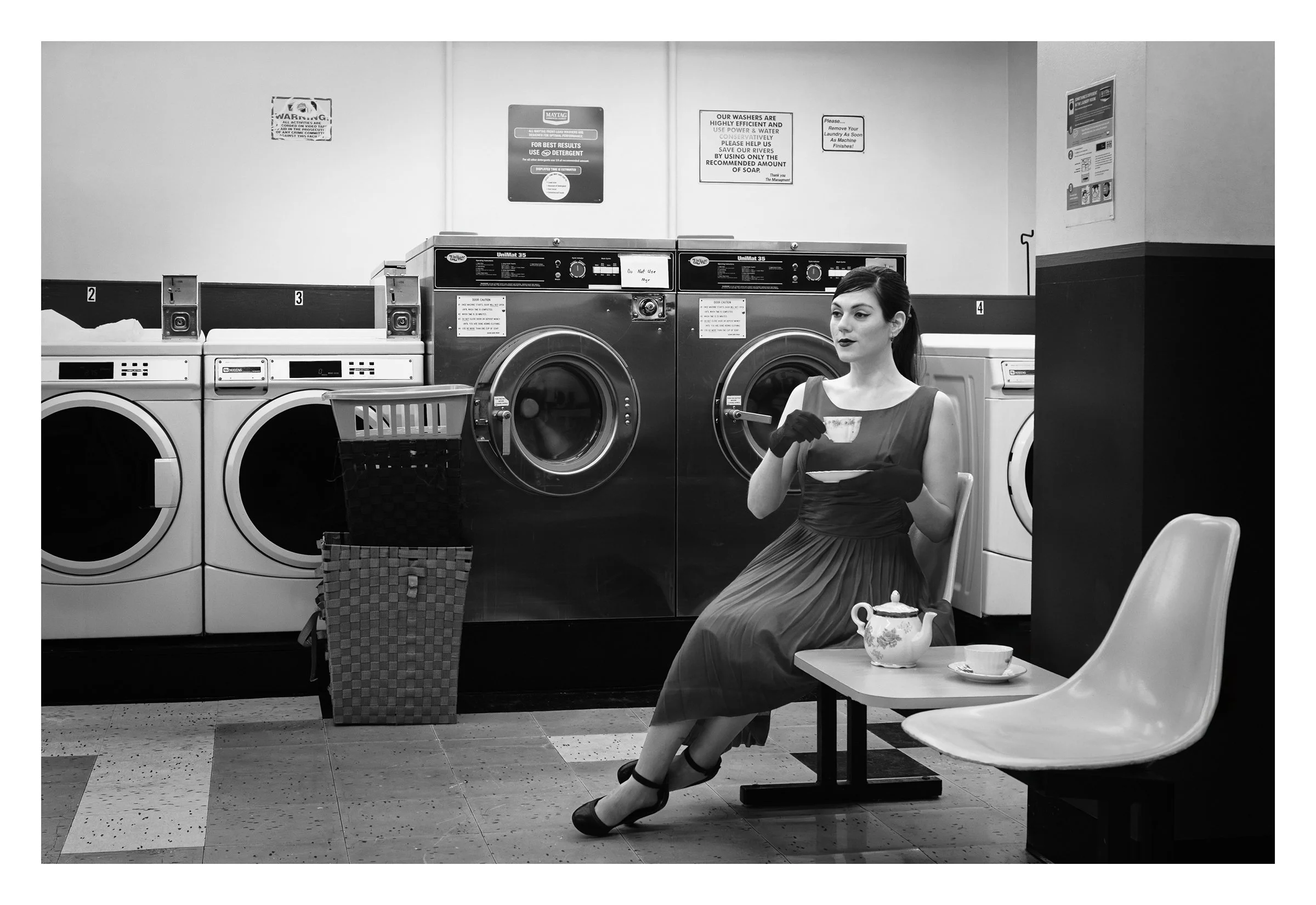 Laundry Time Tea Time, Portland, Oregon, USA 2014 photographed by Dave Haworth. Monochrome photograph of a woman in a formal vintage dress with black gloves sitting in a laundromat, drinking tea from china tea pot and cup while waiting beside washing