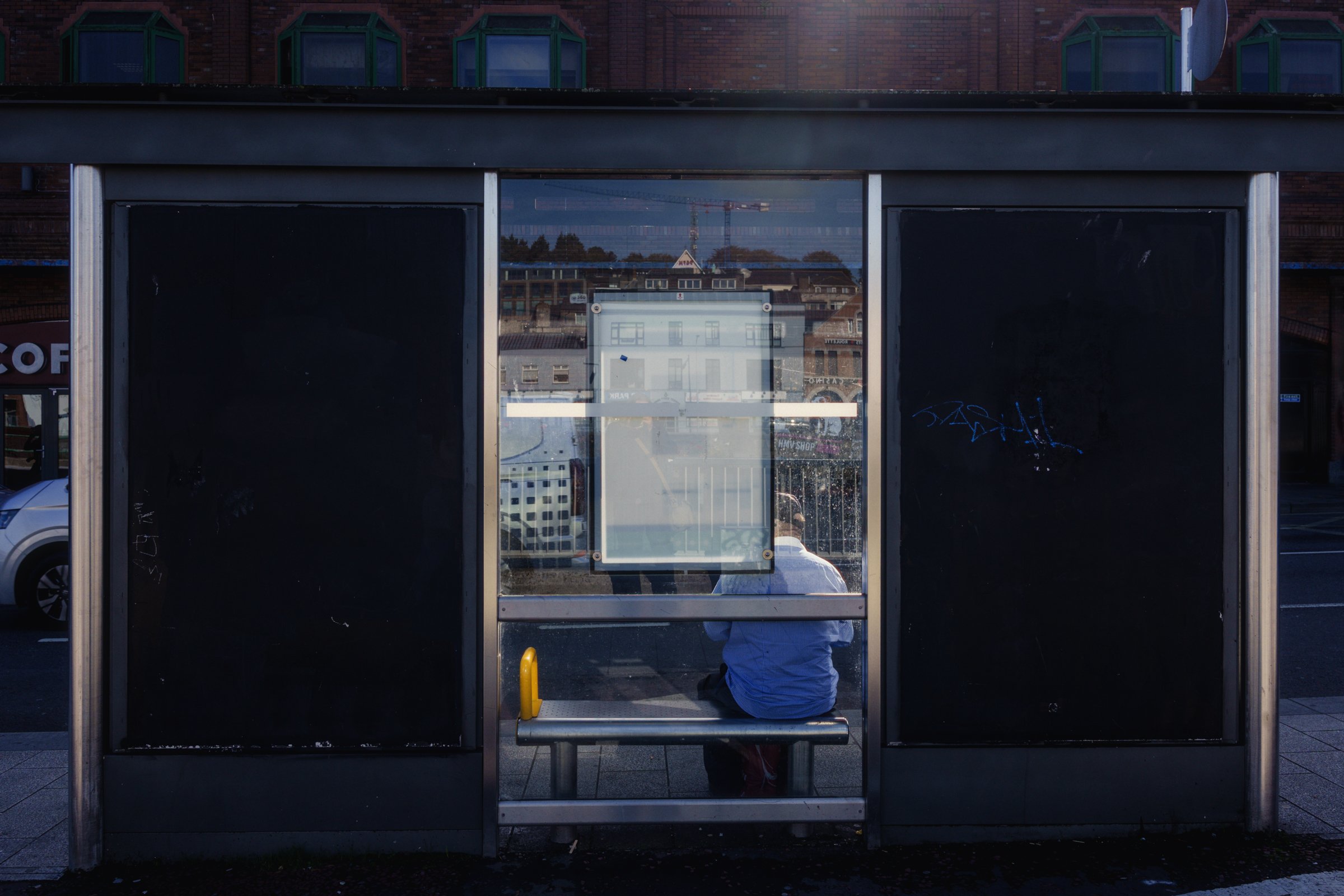 A man sitting at the bus stop in a brilliant ray of sunlight, Cork City, Ireland, 2026 photographed by Dave Haworth