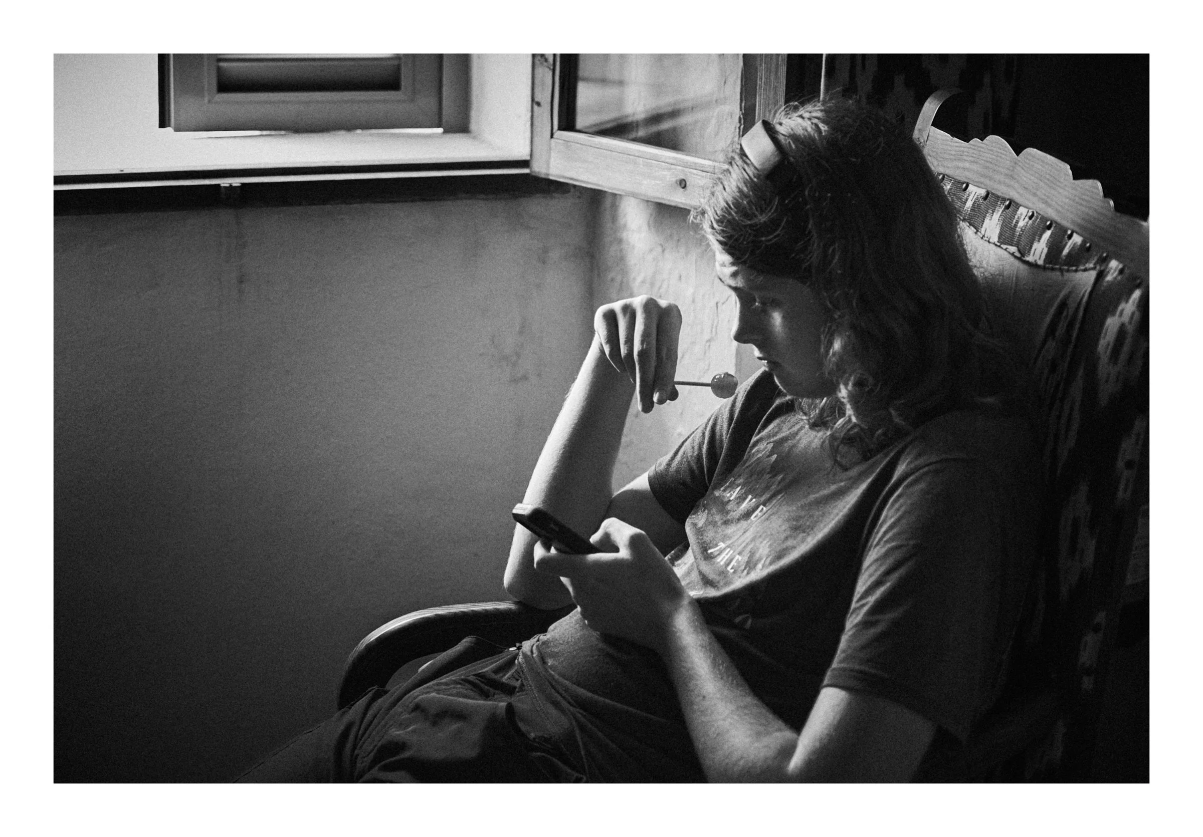 Ian, Mallorca, Spain, 2025 photographed by Dave Haworth. Monochrome photograph of teenage boy sitting by the window in a Spanish Villa looking at this phone.