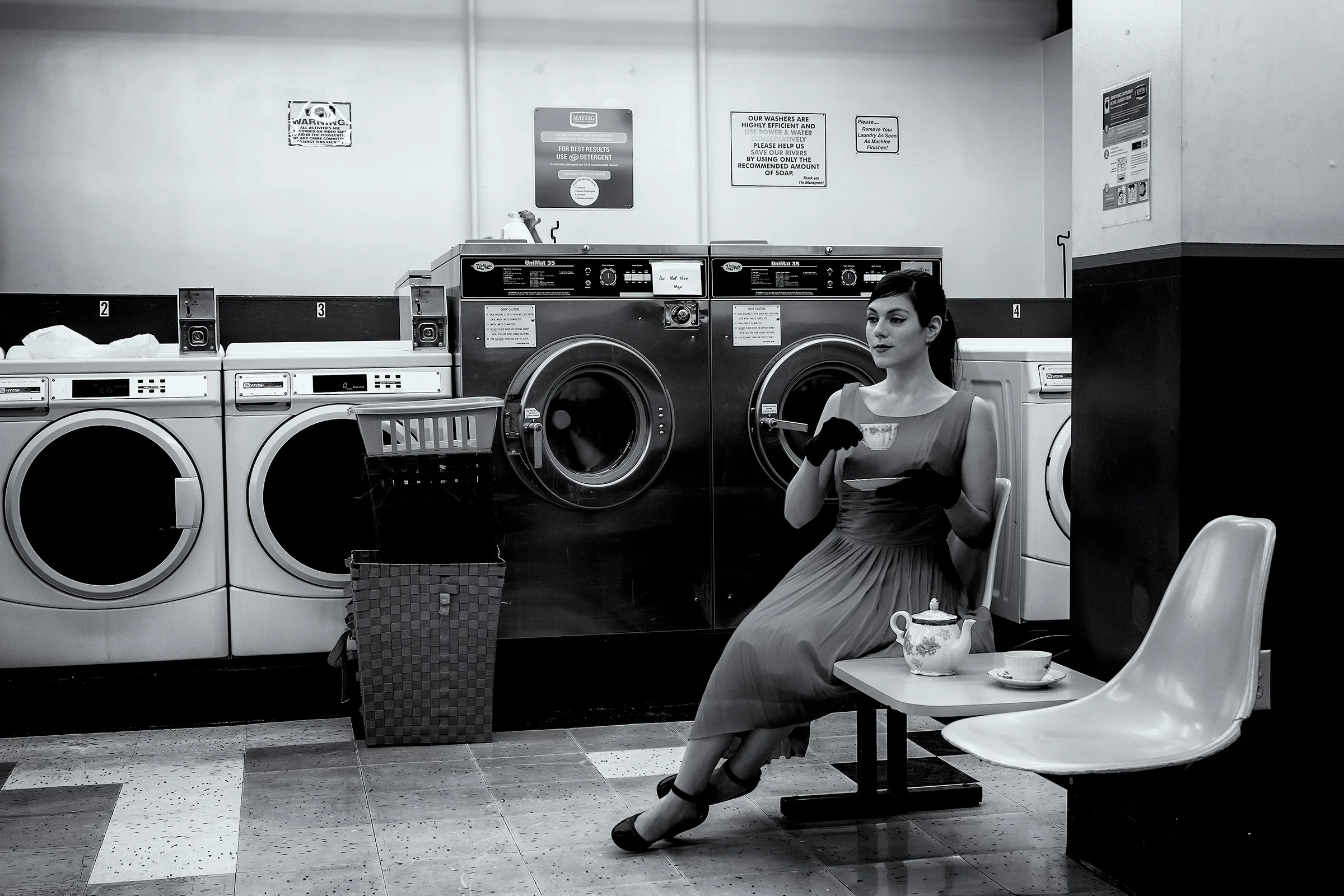 Laundry Time Tea Time, Portland, Oregon, USA 2014 photographed by Dave Haworth. Monochrome photograph of a woman in a formal vintage dress with black gloves sitting in a laundromat, drinking tea from china tea pot and cup while waiting beside washing