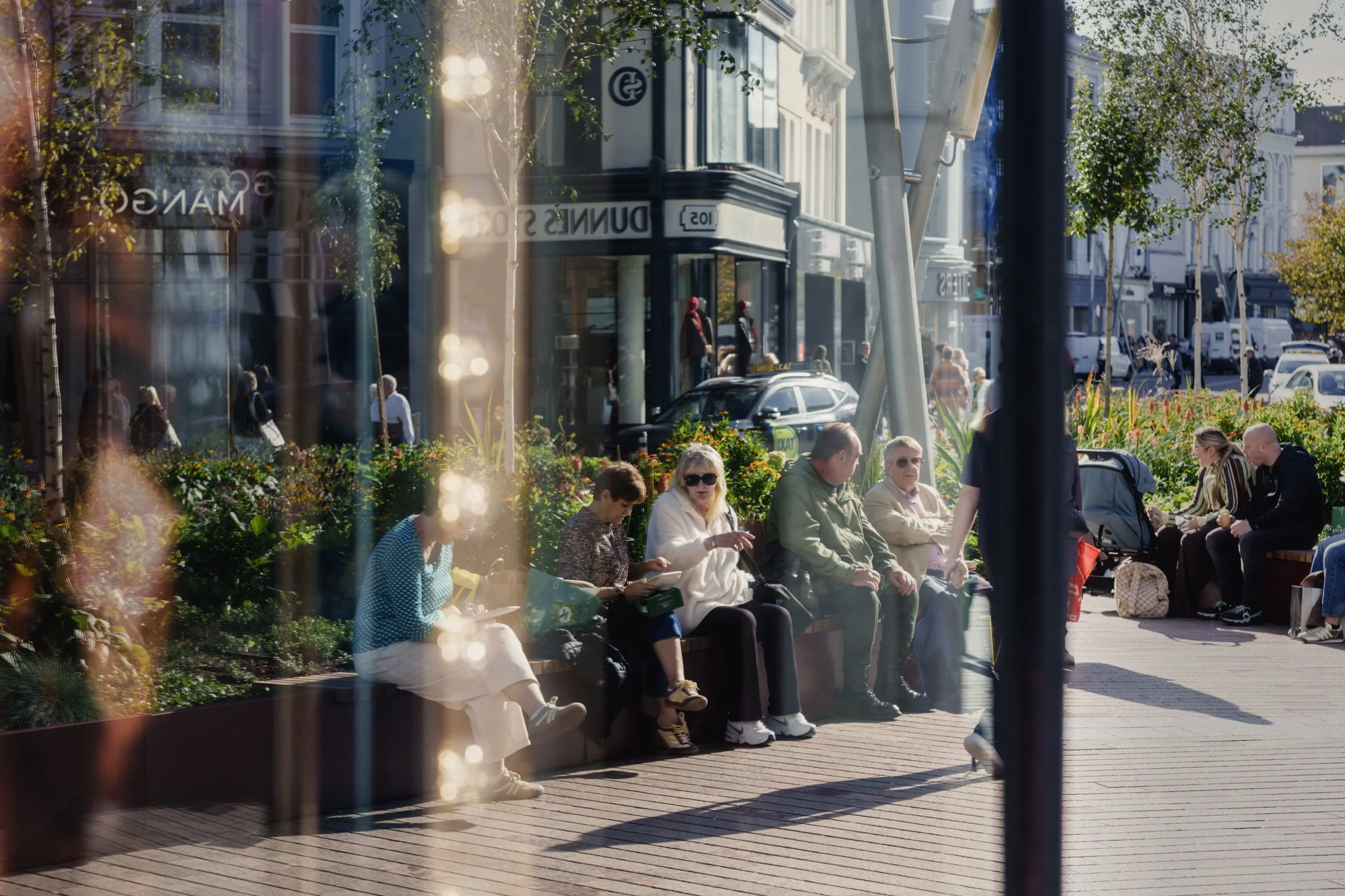 A group of people sitting on the edge of a planter box in Cork City Centre seen through the glass of a shop, Cork City, Ireland, 2026 photographed by Dave Haworth