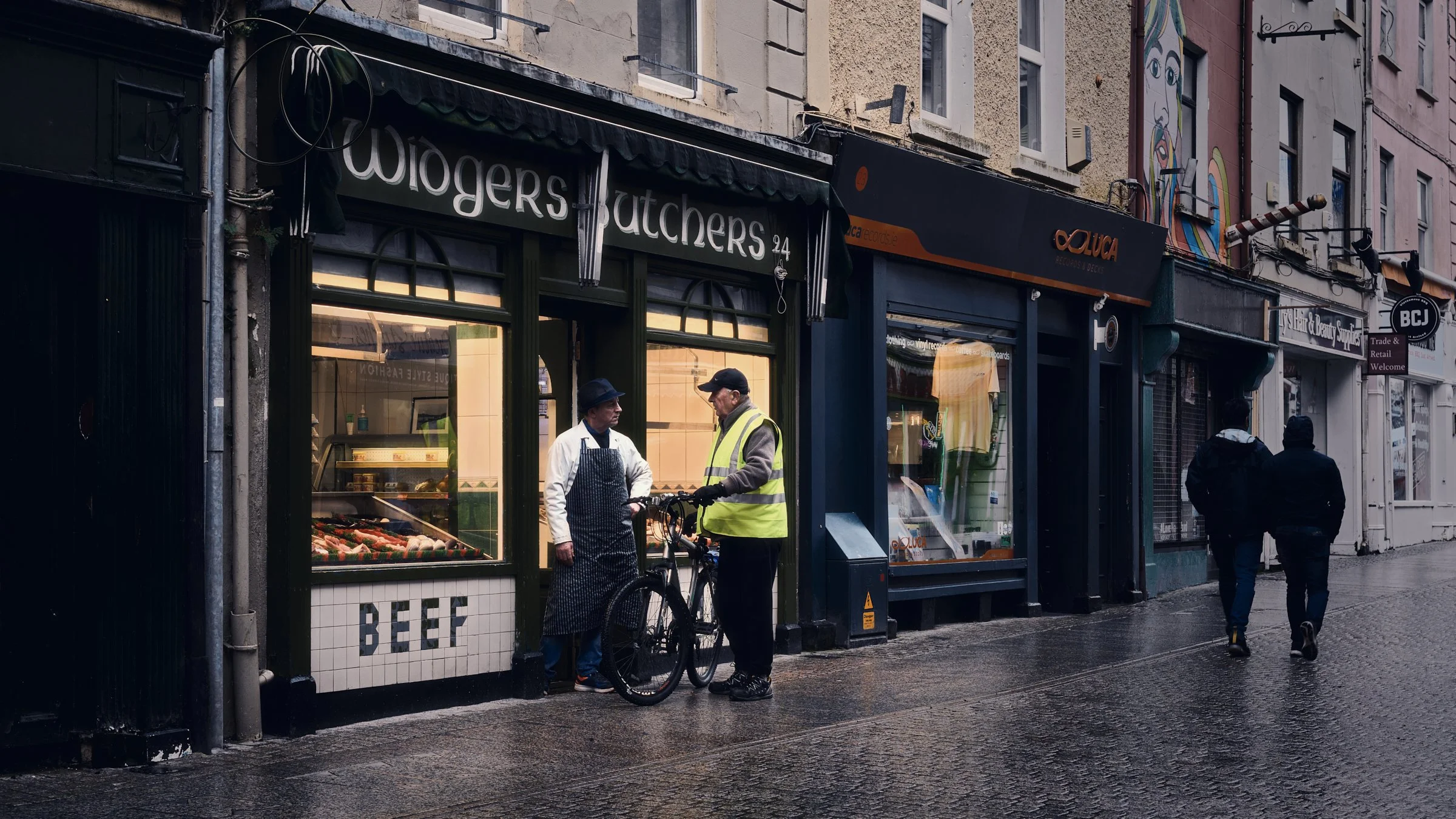 One Take - Ireland "The Butcher", Waterford City, Co. Waterford, Ireland 2024. A cinematic color photograph of the Butcher & a man with the bike chat in front of Widgers Butchers in Waterford City Centre.