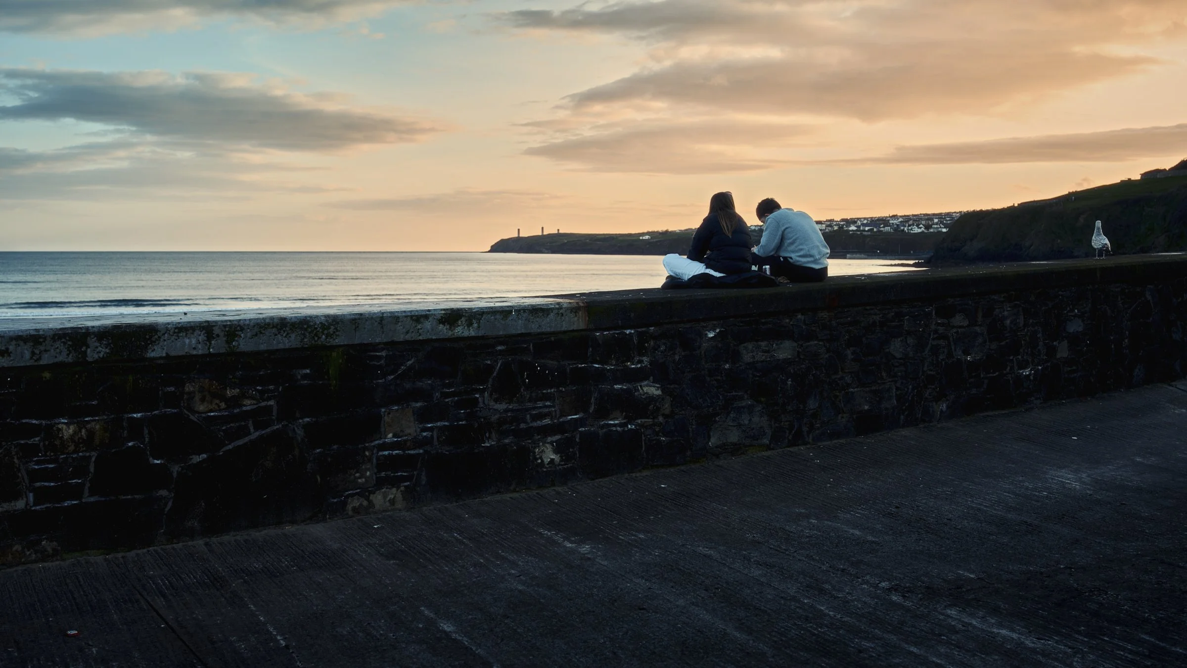 One Take - Ireland "On Edge", Tramore, Co Waterford, Ireland 2026. Cinematic color photograph of a young couple sitting on the sea wall chatting at sunset, with the Tramore Metal Men in the distance.