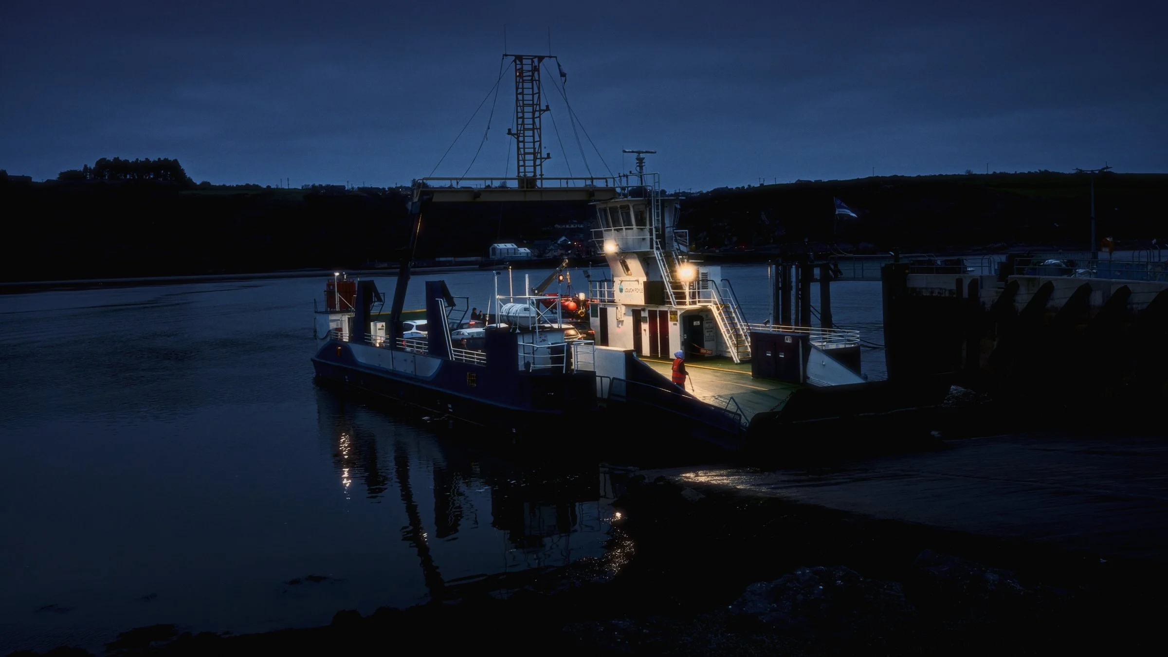 One Take - Ireland "The Ferryman", Passage East, Co. Waterford, Ireland 2025. A cinematic color photograph at  dusk view of the Passage East Ferry lit up with a crew member lit by the conning tower lights.