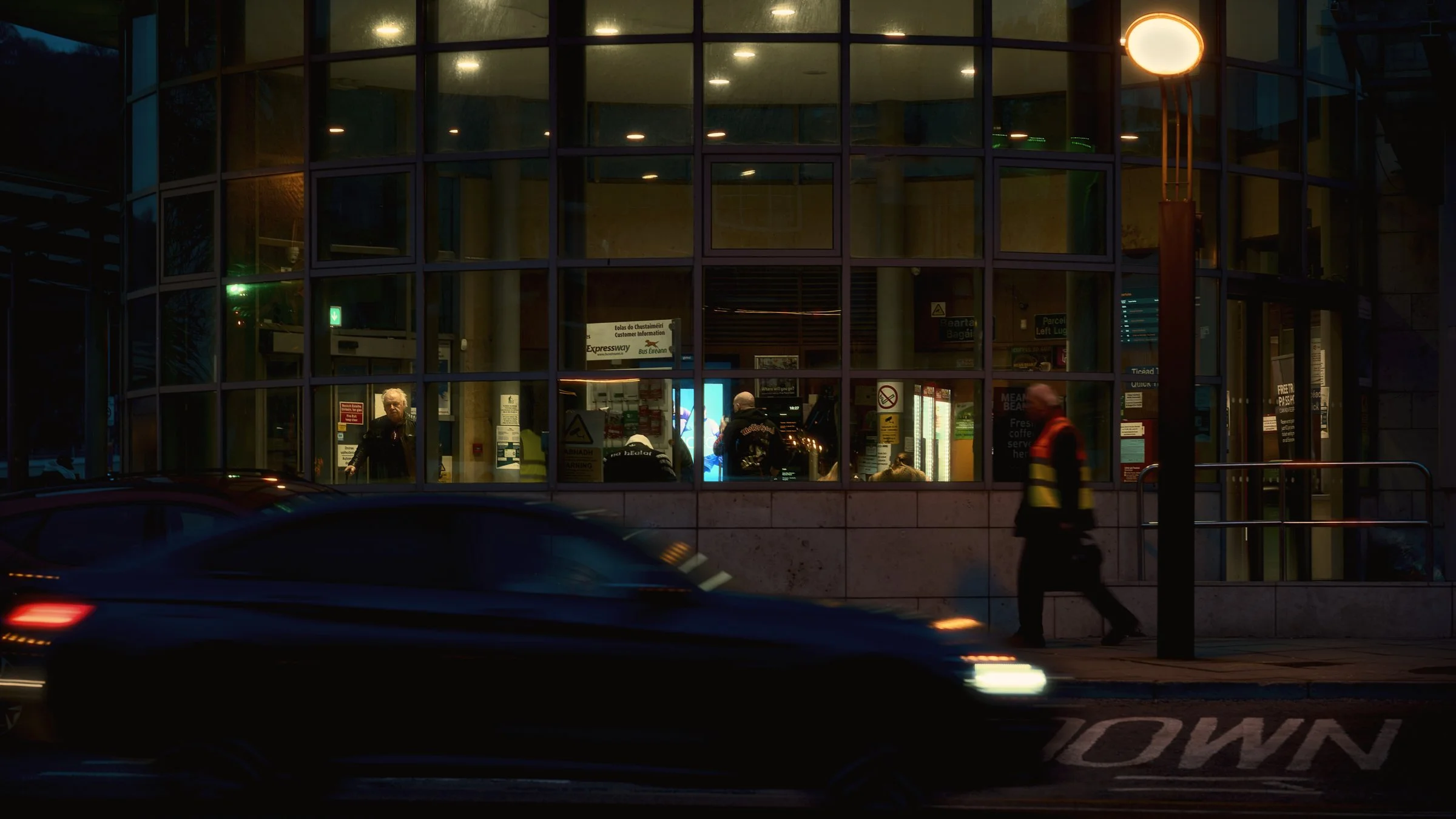 One Take - Ireland "Waiting", Waterford Bus Station, Ireland 2026. A cinematic color photograph of Waterford bus station at night looking in the lit interior of the bus station from outside. A man is looking out towards the camera from inside.