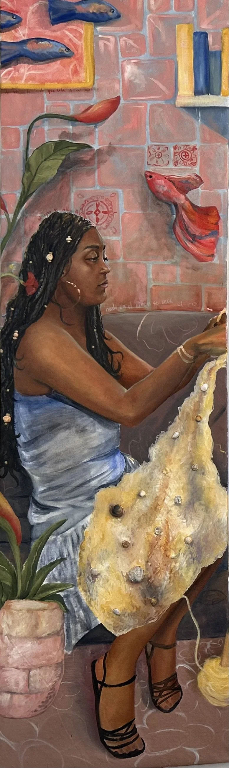 A woman with dark braided hair adorned with shells, wearing a blue dress, is sitting and looking at her phone, surrounded by colorful paintings of fish, a potted plant, and decorative tiles on a pink brick wall.