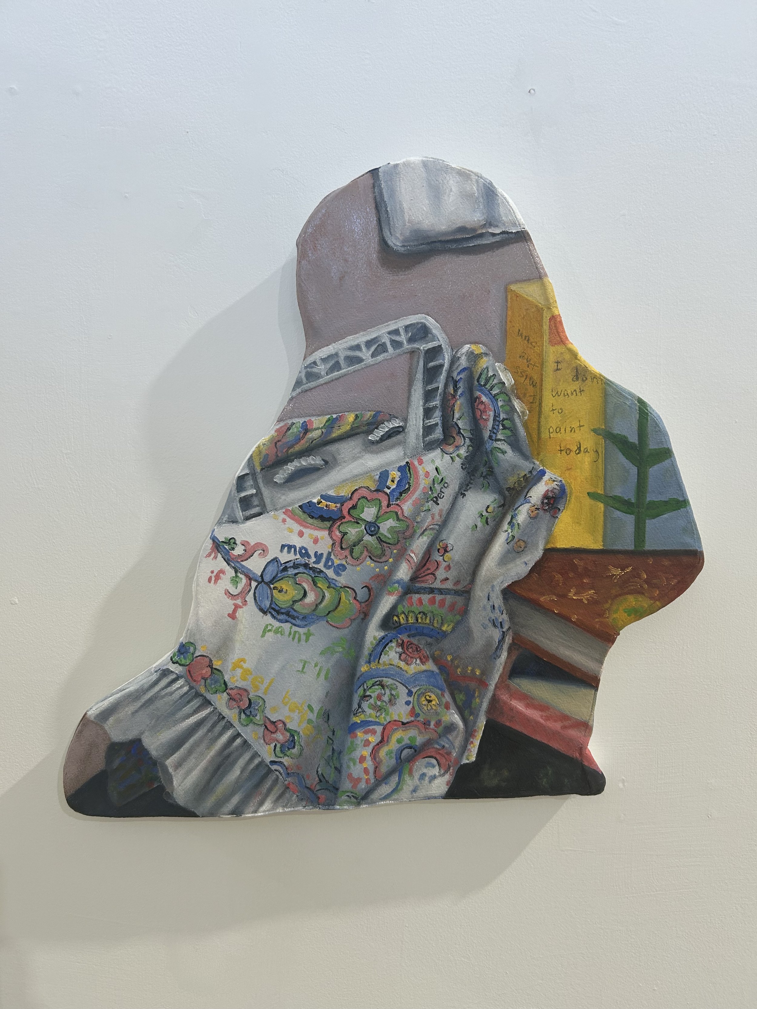 Painted cutout of a person wearing a hoodie with colorful embroidery, sitting on a stool, with a yellow book to their side that has handwritten text.