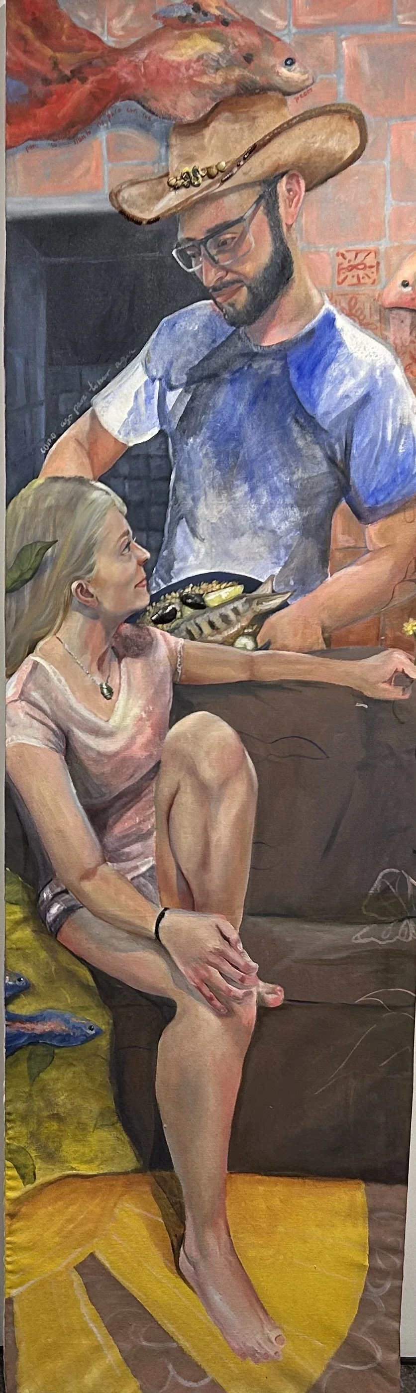 A man in glasses and a woman with a leaf in her hair sitting on a couch, with the man holding a plate of fish and shellfish, and the woman looking up at him, both in a cozy indoor setting.