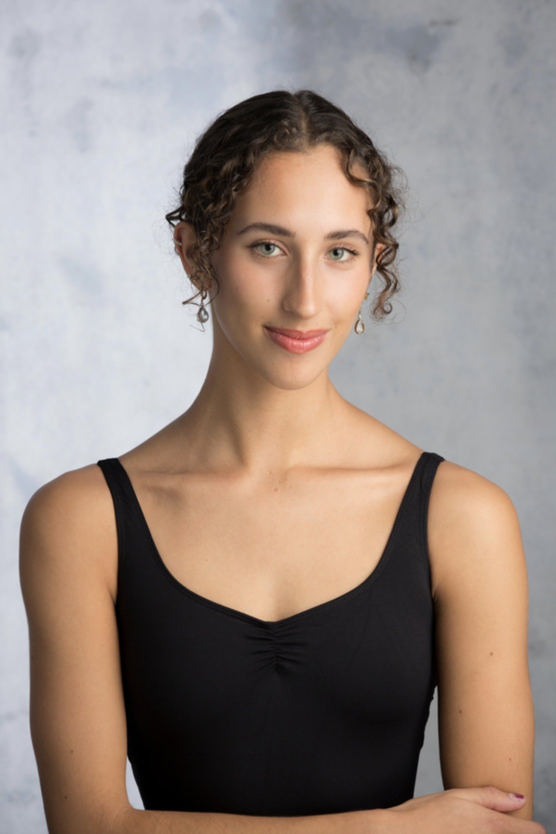 A young woman with curly brown hair and blue eyes, wearing a black tank top and earrings, smiling softly in front of a plain gray background.
