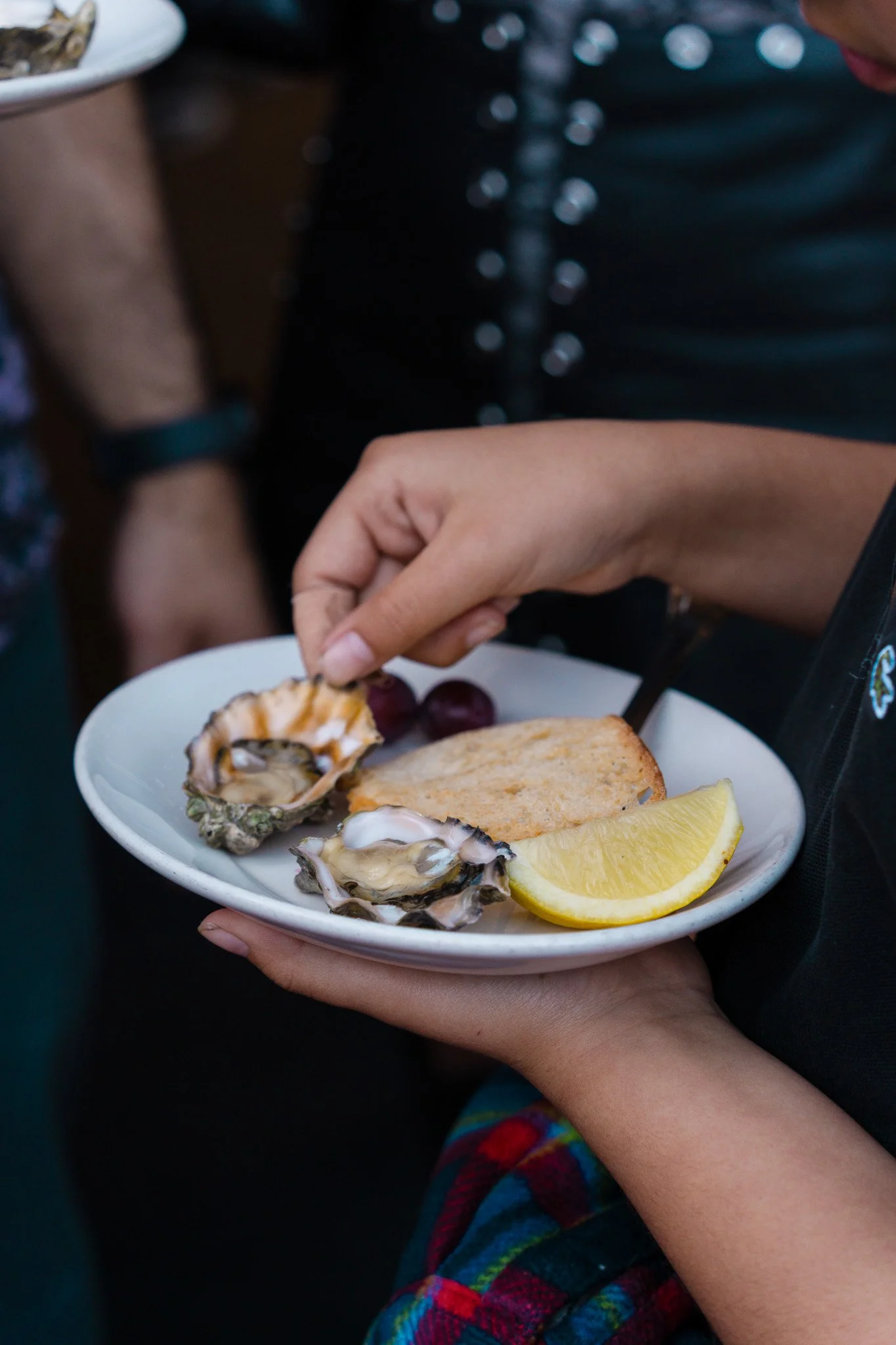 A person holding a white plate with oysters, a lemon wedge, two grapes, and a piece of toast at a community gathering in San Francisco, California. 