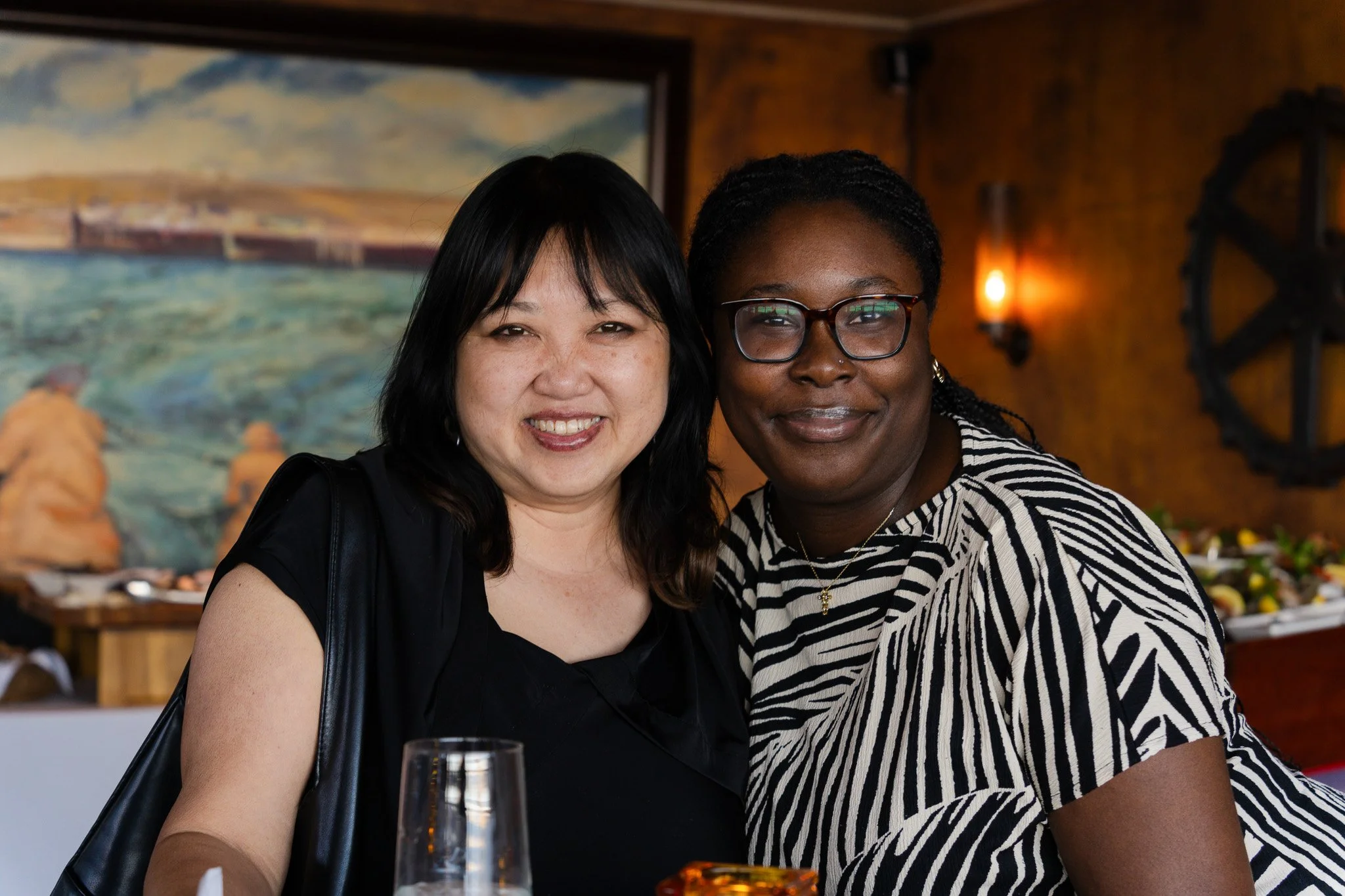 Two women smiling and posing together indoors during a social gathering or meal, with a table of food and drinks in the background.