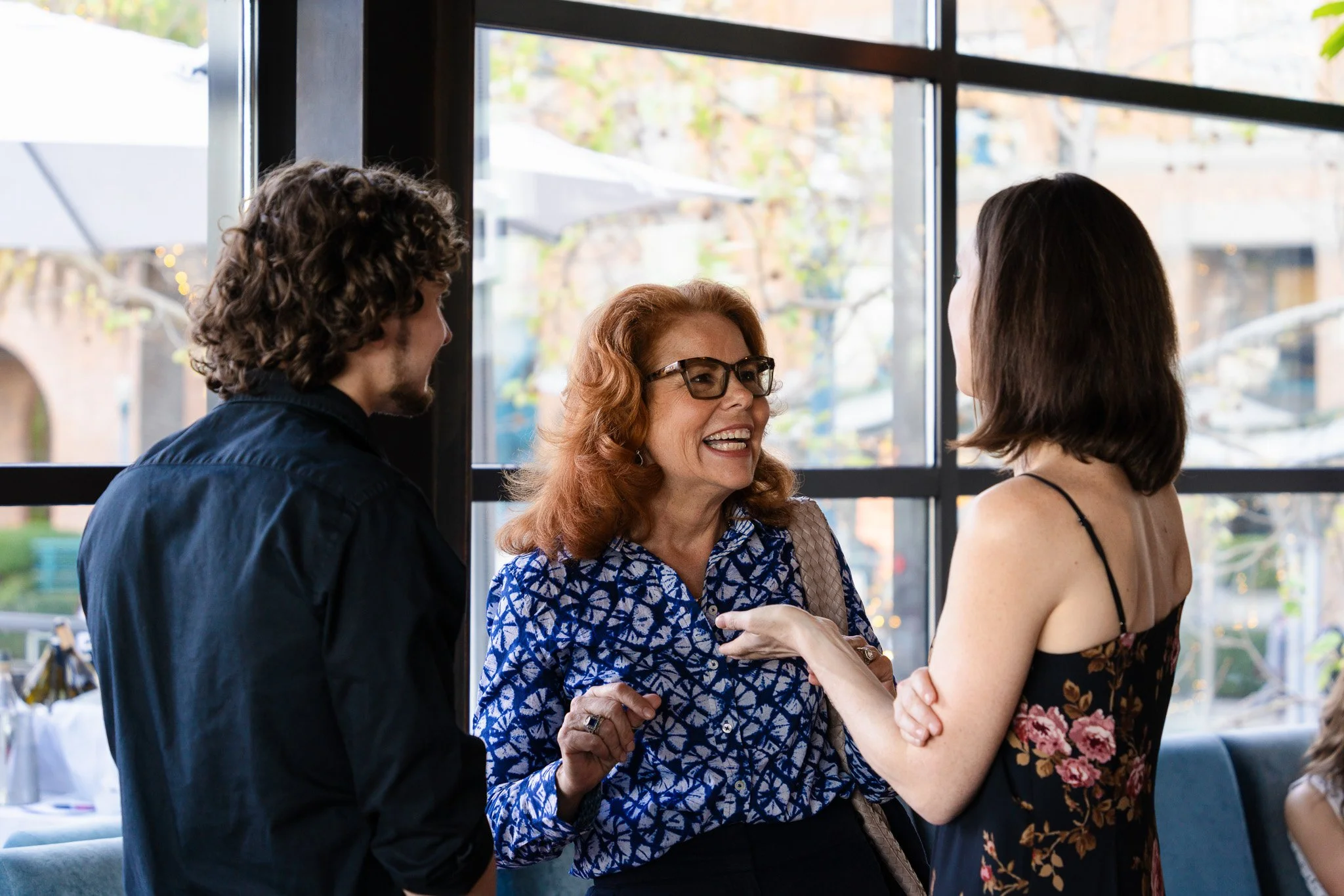 Three people talking and smiling inside a cafe with large windows showing trees outside at the waterfront in San Francisco, California.