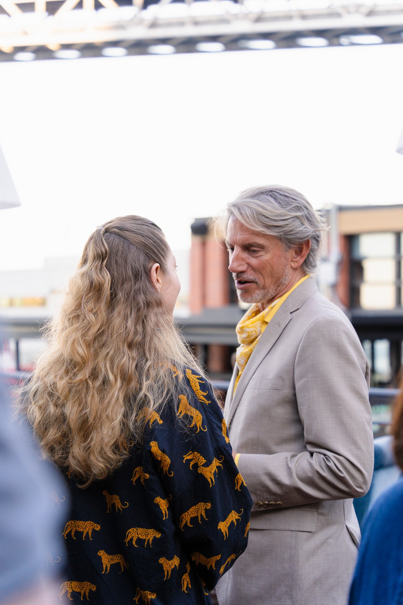 A man and a woman engaged in conversation, outdoors, with buildings in the background. The woman has long, wavy hair and wears a dark jacket with a tiger print. The man has gray hair, wears a light-colored suit, and a yellow tie. 