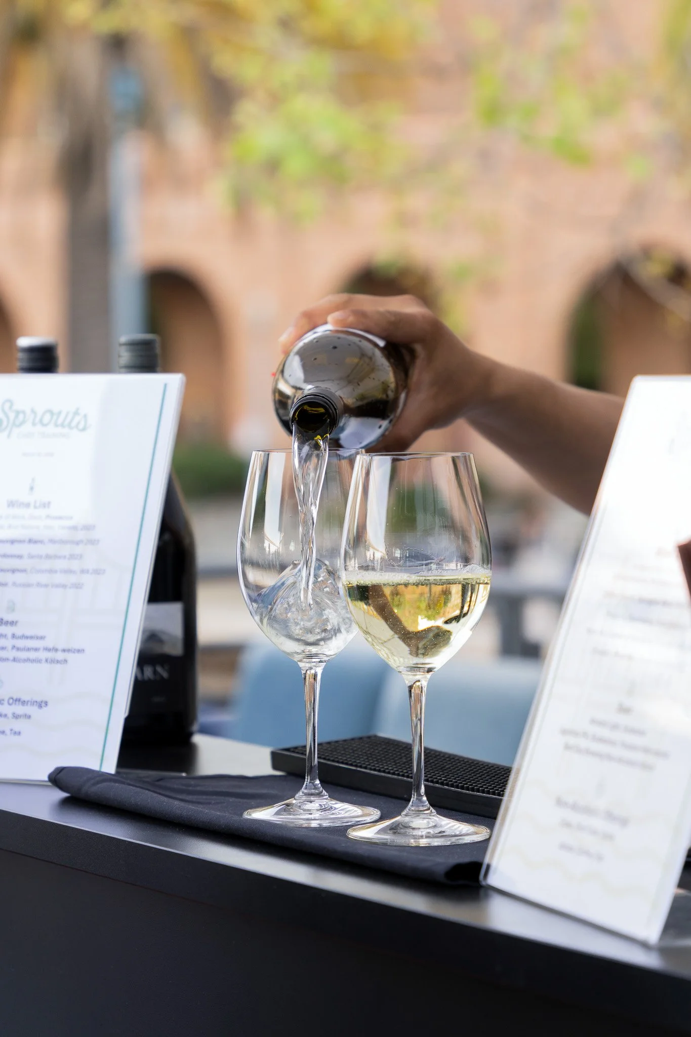 A person is pouring white wine into a wine glass at an outdoor restaurant table with menus and bottles present, taken at an outdoor event in San Francisco, California.