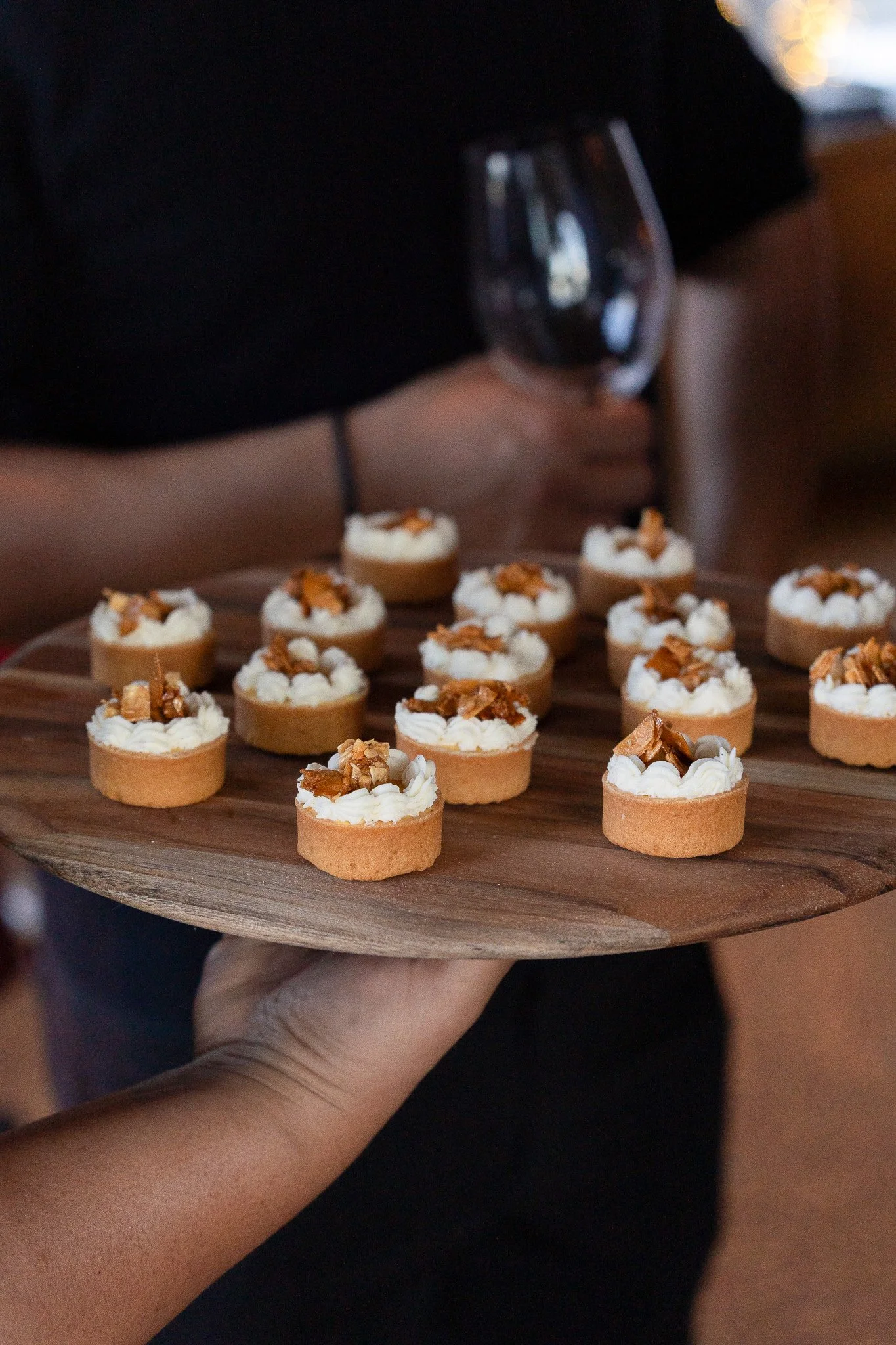 A hand holding a wooden tray with small tartlets topped with whipped cream and caramelized nuts, with a person in the background holding a glass of red wine, taken at a community event in San Francisco that features food and restaurants.