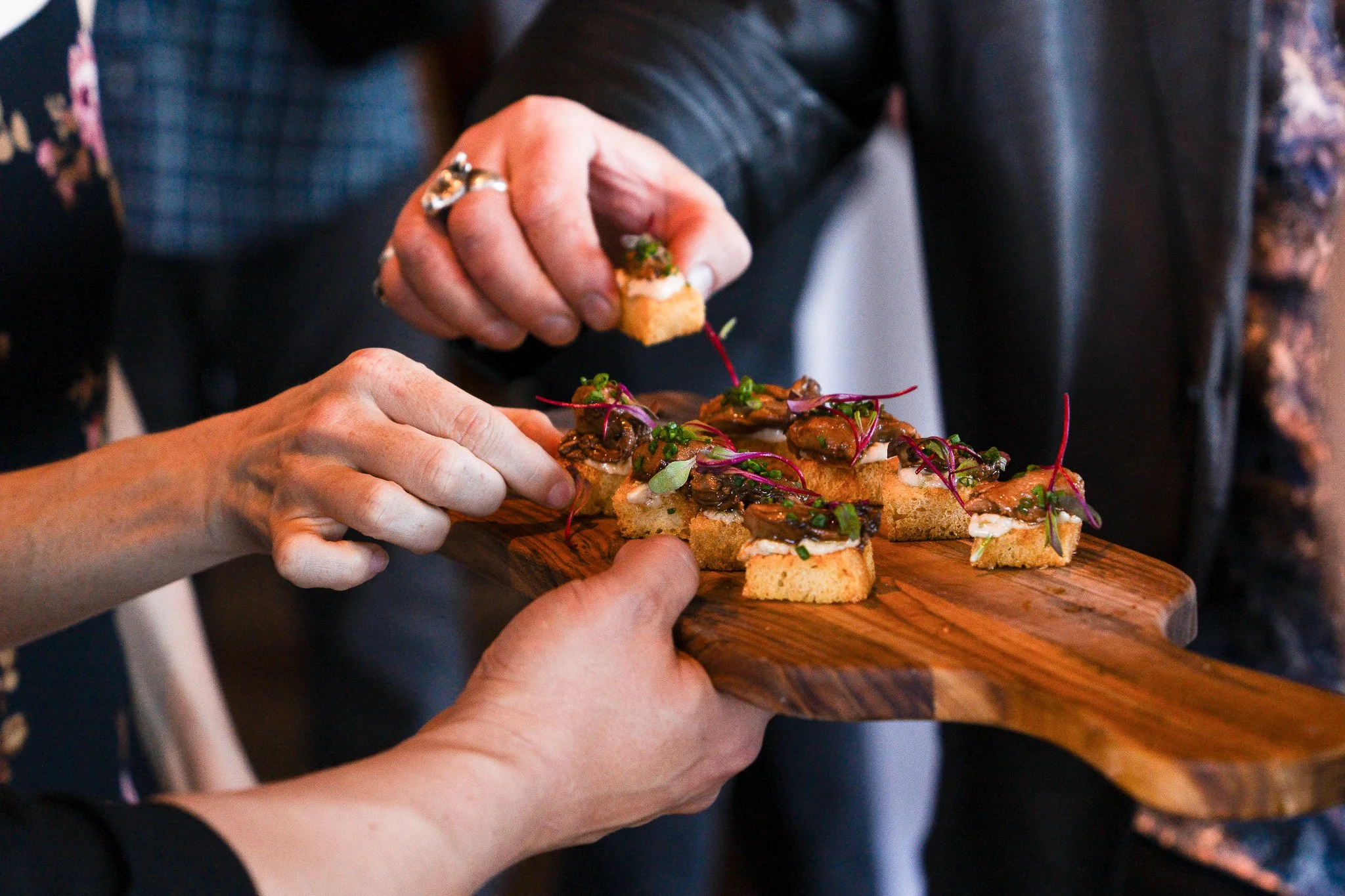 Close-up of two people’s hands serving and receiving a wooden platter of gourmet appetizers topped with microgreens and sauce at a community event.