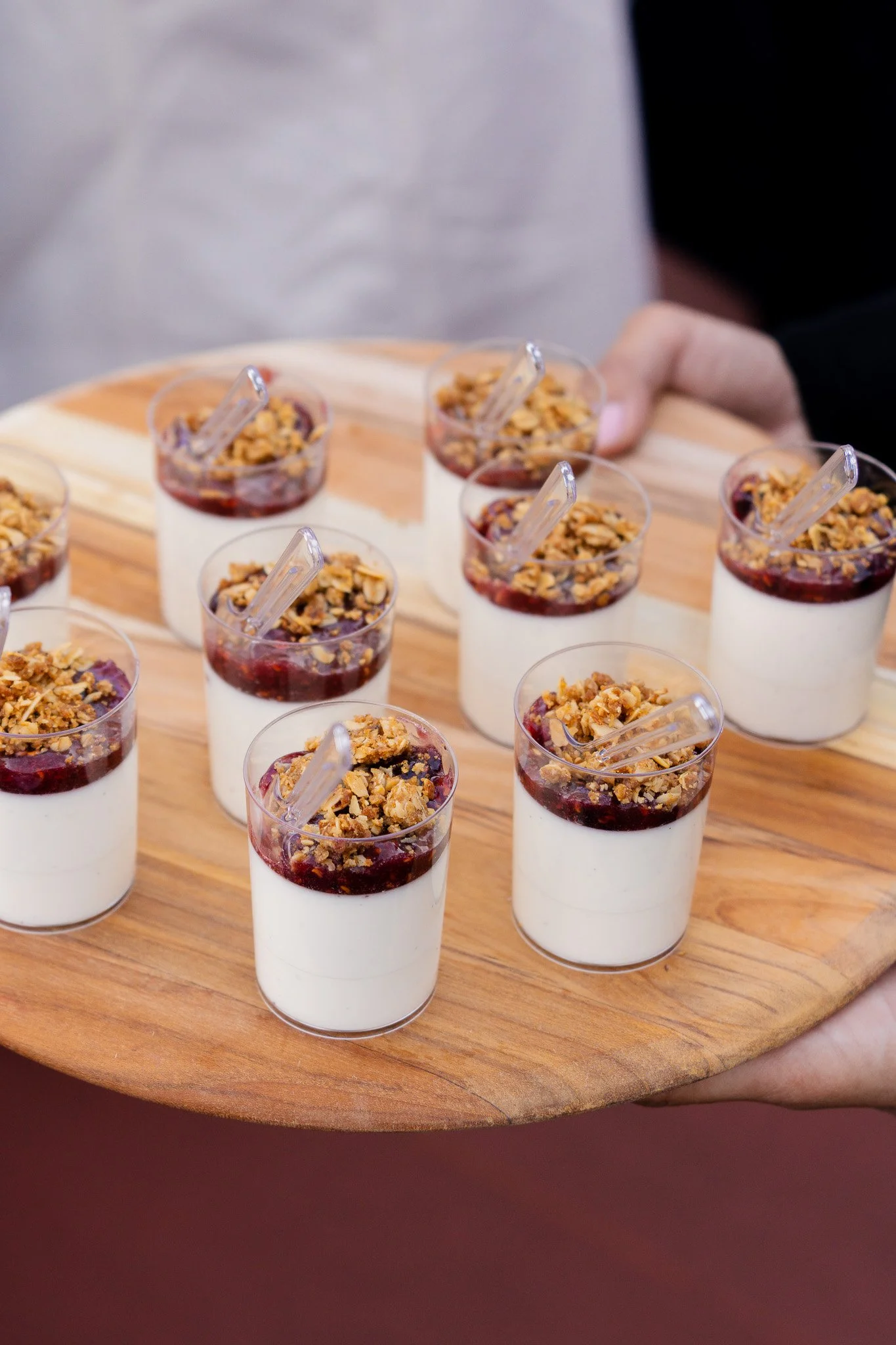 Multiple small cups of layered dessert with white cream, berry jam, and granola on a round wooden tray being held by a person at an outdoor party in San Francisco, California. 