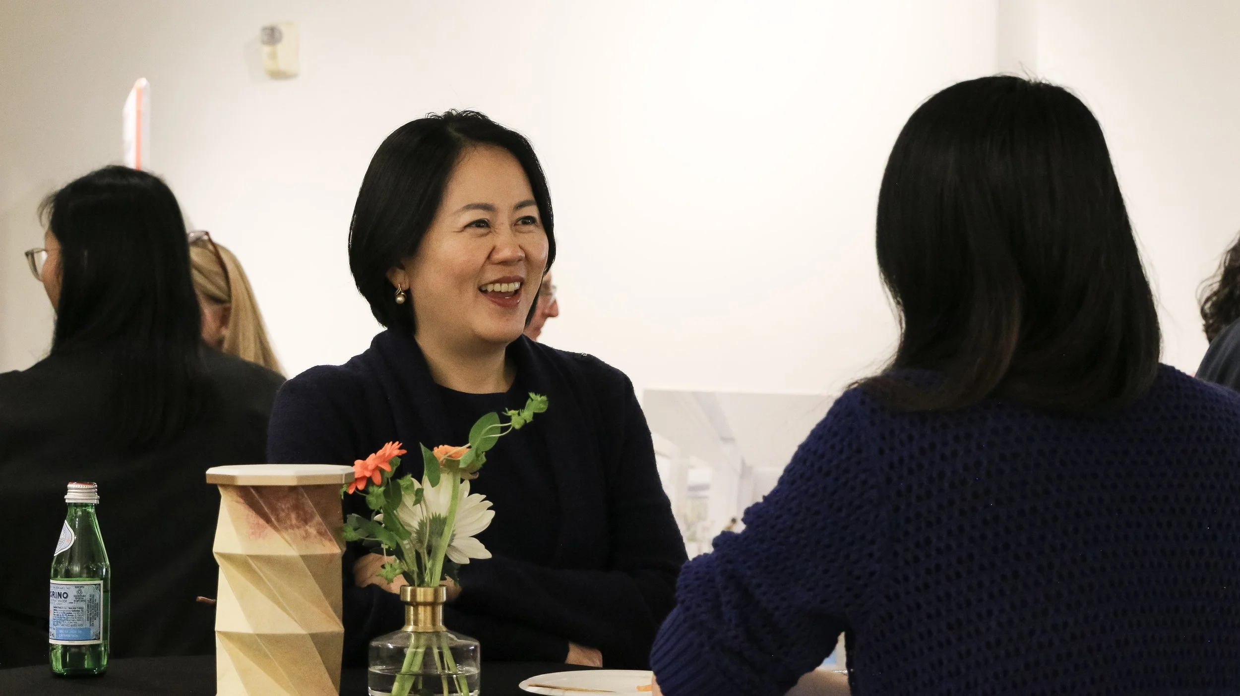Two women sitting at a table, engaged in conversation, with a decorative vase and a small water bottle on the table. The woman on the left has short black hair and is smiling, while the woman on the right has long dark hair and is facing away from th