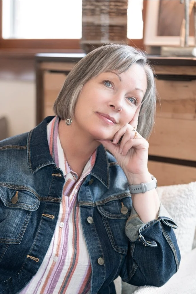 A woman with short gray hair and blue eyes, wearing a striped shirt and denim jacket, resting her head on her hand in a cozy indoor setting.