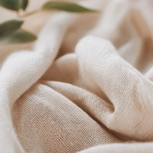 Close-up of beige fabric with soft folds and a few green leaves in the background.