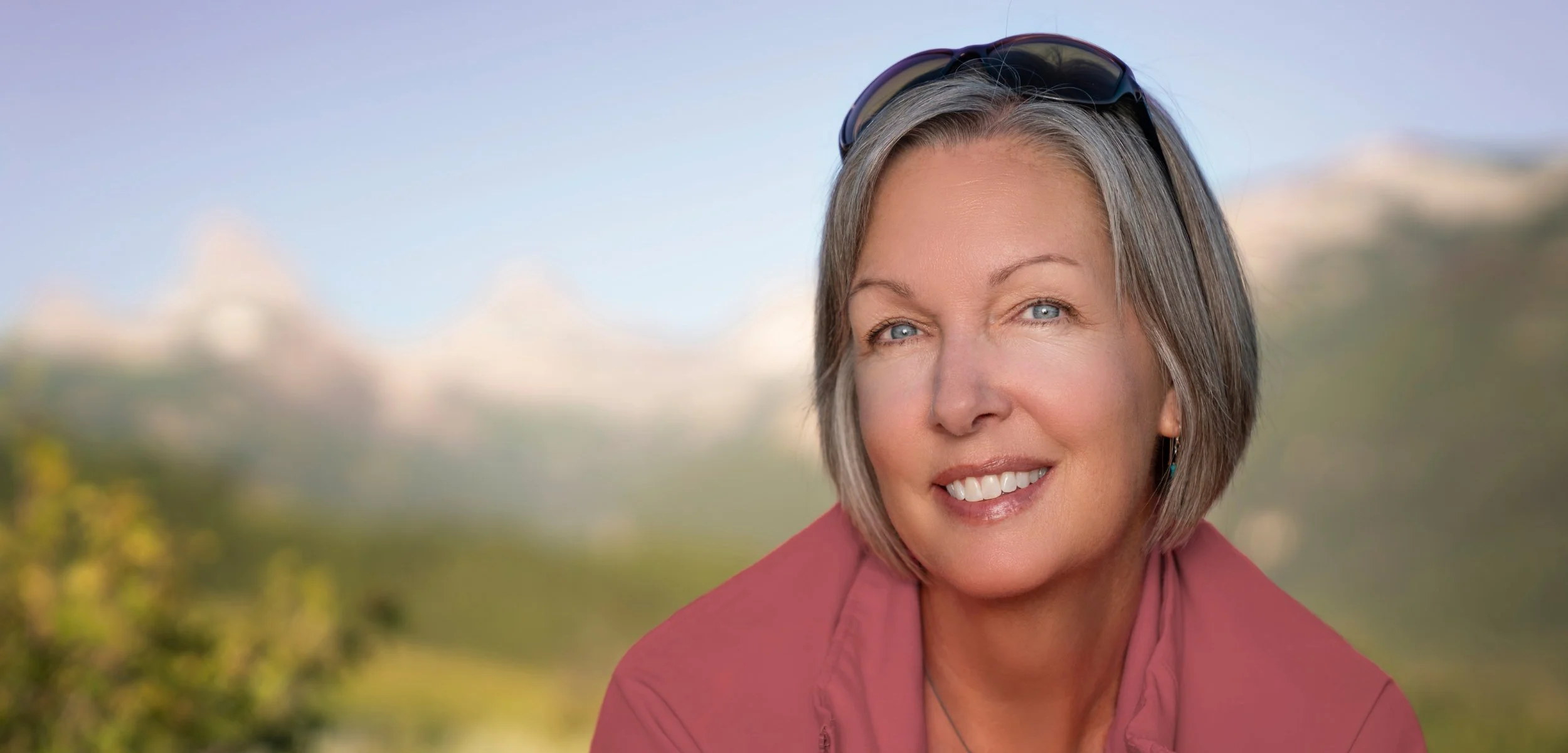 A smiling woman with short gray hair, wearing sunglasses on her head and a pink jacket, outdoors with mountains in the background.