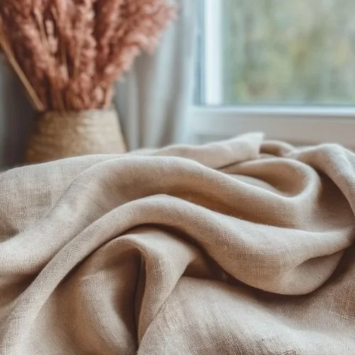 Close-up of beige fabric draped on a surface with a window and a vase with pink dried flowers in the background.