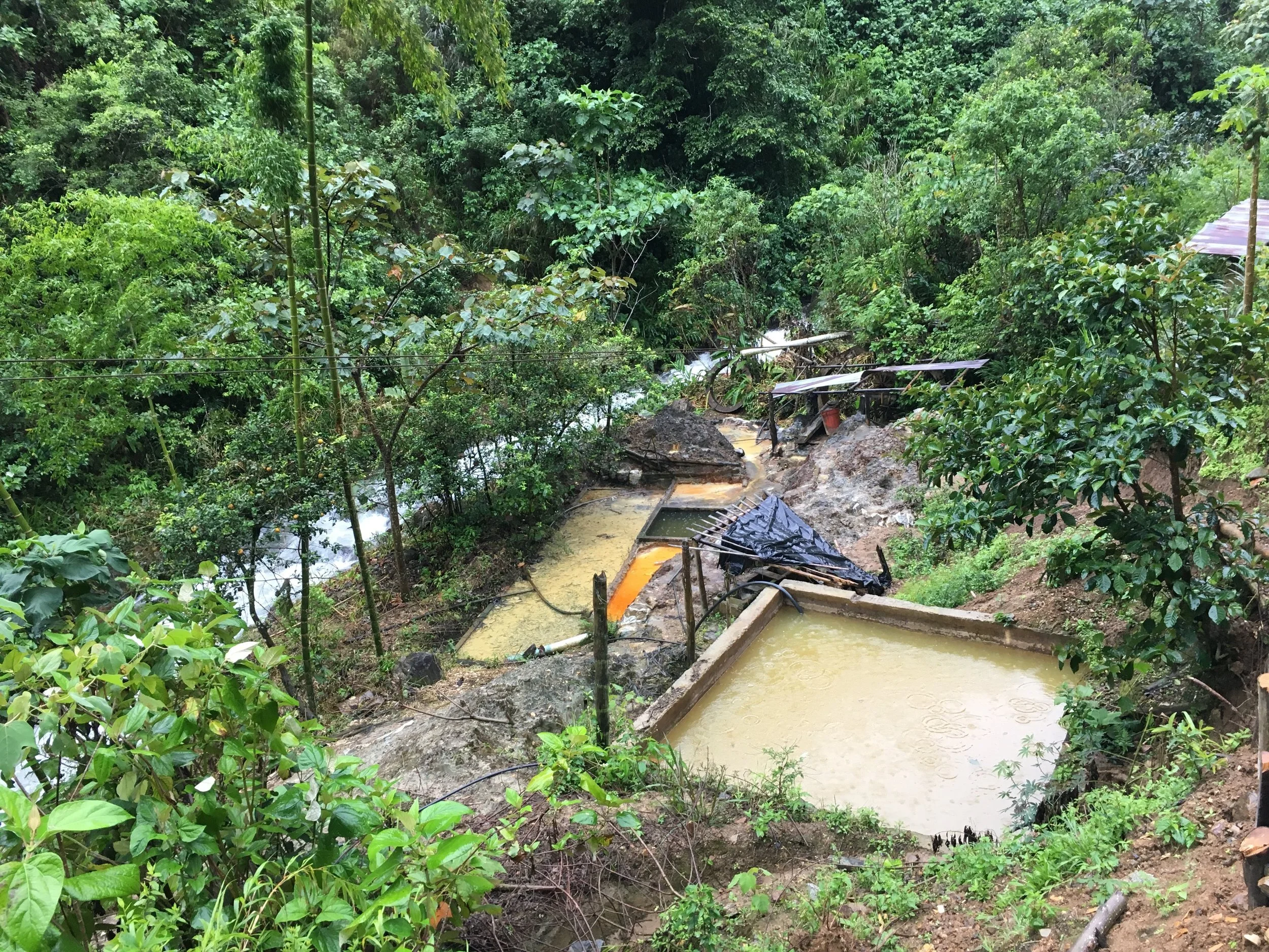 Erosion on a hillside with small pools of water, surrounded by dense green forest and some makeshift structures.